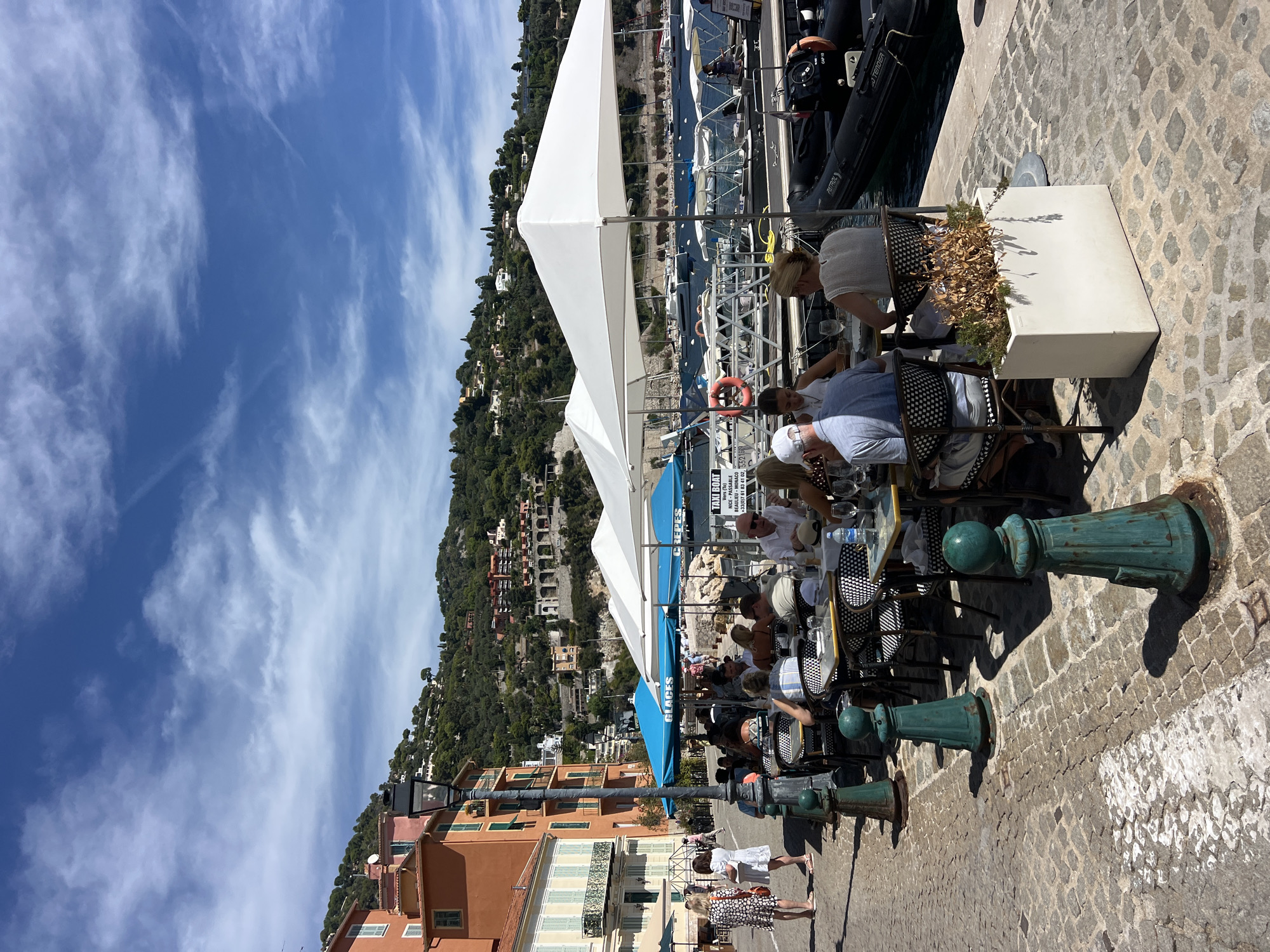 The image depicts a waterfront scene in Villefranche-sur-Mer, France, on a sunny day with scattered clouds. The foreground is a cobbled stone walkway, lined with teal bollards. To the left, a row of buildings displays characteristic Mediterranean architecture with orange and ochre facades, green shutters, and balconies. Several people are walking on this walkway.
Dominating the mid-ground is an outdoor seating area for a restaurant, shaded by large white and blue umbrellas. Tables are set with checkered cloths, and diners are seated, engaged in conversation and eating. The blue umbrellas are labeled "GLACES" and "CREPES".
Beyond the restaurant, a harbor is visible, filled with various boats, including yachts and smaller vessels docked along a pier. Some of these boats appear to be tender boats. A sign on a white structure near the boats advertises "TAXI BOAT" with contact information. Inflatable boats are also present along the waterfront.
The background features a steep, verdant hillside densely populated with buildings, characteristic of the French Riviera. The overall impression is of a lively, upscale tourist destination.