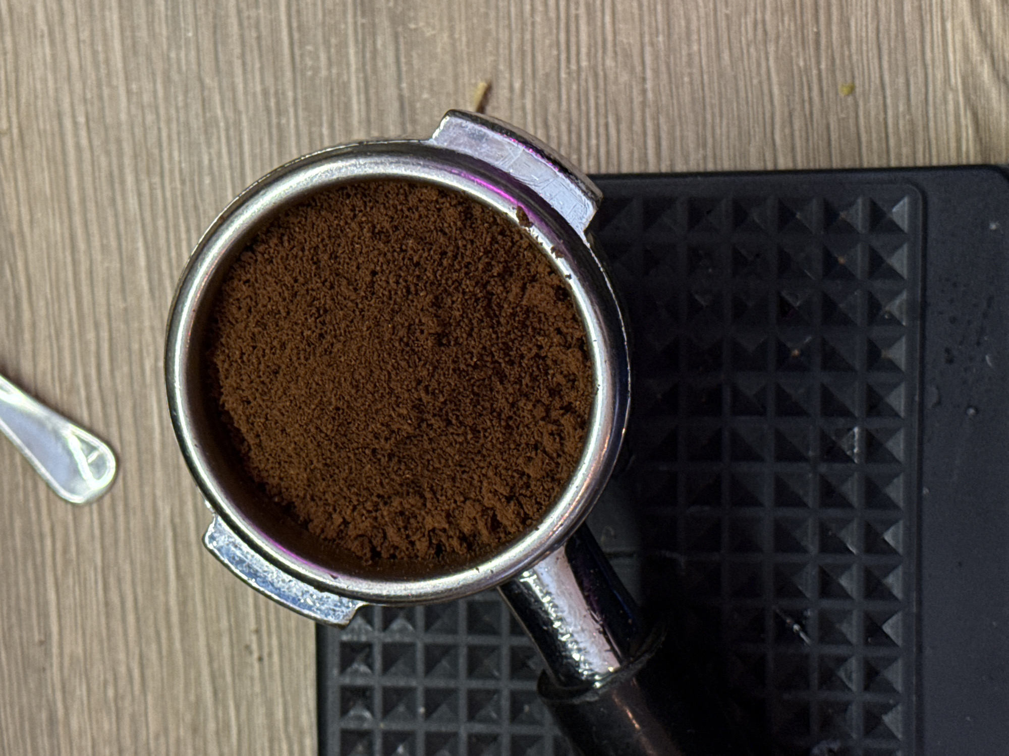 The image shows a close-up, overhead view of a portafilter filled with finely ground coffee. The portafilter is resting on a textured black surface, which appears to be a tamping mat. The background is a wooden surface with a prominent grain. A shiny metal spoon is partially visible in the upper portion of the image. The lighting suggests an indoor setting, possibly a kitchen or cafe. There are no people or animals present, and no specific geographical markers are visible to indicate a location in Moscow, Russia.