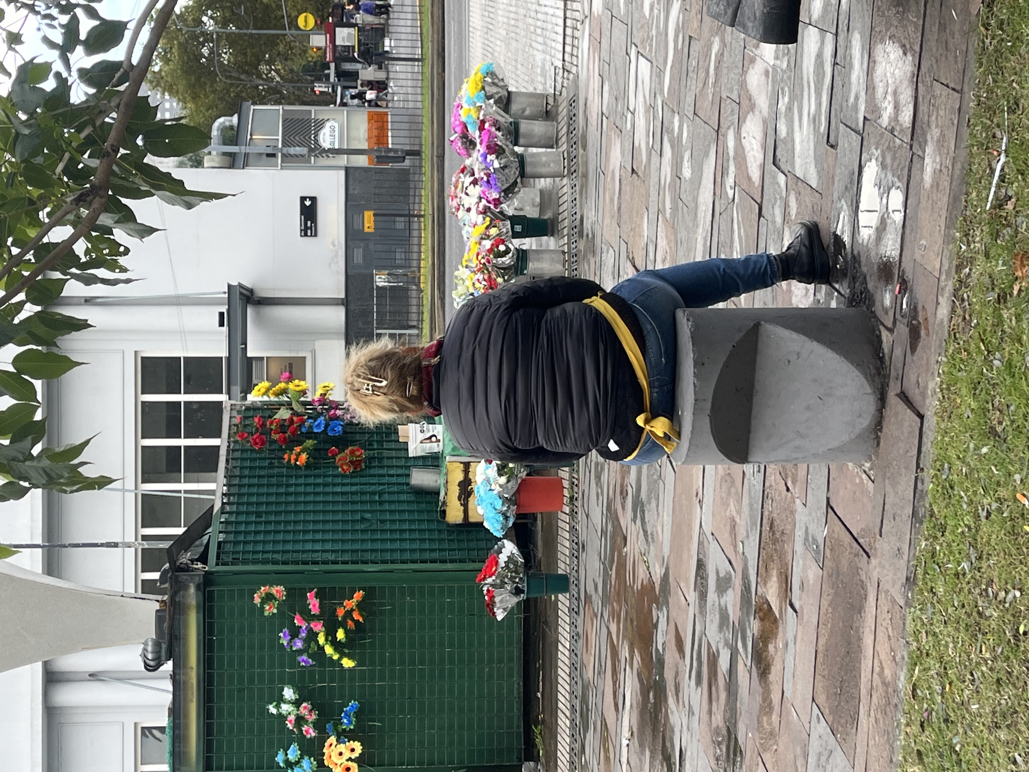 A woman sits on a concrete cylinder next to a green metal fence adorned with artificial flowers in Buenos Aires, Argentina. The woman wears a black puffer jacket with a yellow strap, blue jeans, and black boots. A line of flower arrangements in metallic containers sits behind her. The location appears to be an urban public space with brick pavement and grass. Buildings and street signs are visible in the background.