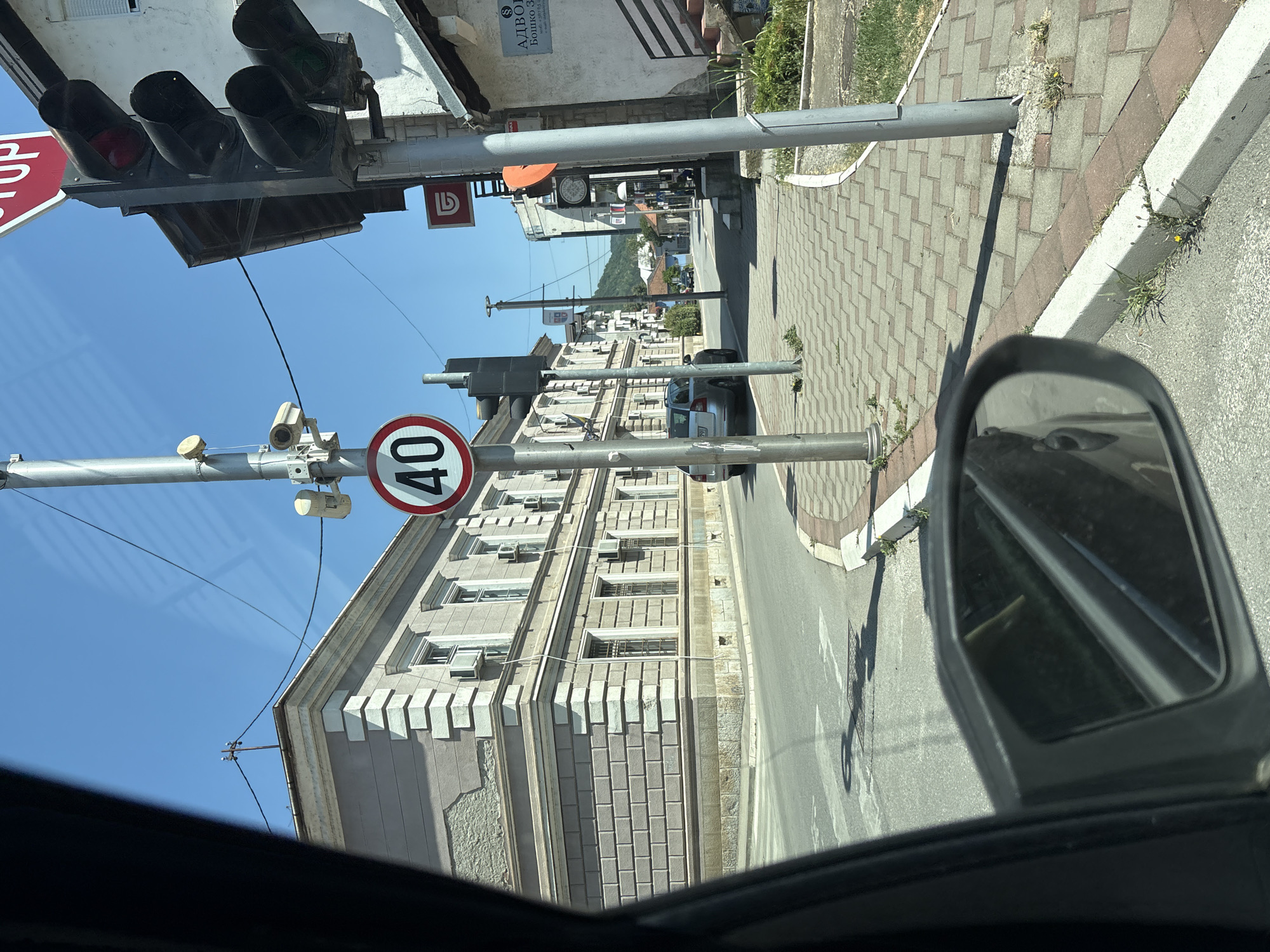 The image depicts a street scene in the city of Zvornik, Bosnia and Herzegovina, viewed from inside a vehicle. The perspective is primarily from the driver's seat, as indicated by the presence of a side mirror in the lower left of the frame.
In the foreground, the road surface is visible, with a pedestrian crossing marked by white lines. A gray sedan is parked on the right side of the road, facing away from the viewer. Adjacent to the road is a paved sidewalk made of interlocking bricks.
Dominating the center of the image is a utility pole with various fixtures attached. These include a traffic light with red and green signals illuminated, a speed limit sign indicating "40" within a red circle, and what appear to be security cameras. Further down the street, another utility pole is visible.
On the left side of the street, a large, ornate building with classical architectural elements stands prominently. It has multiple windows and air conditioning units installed. The building's facade is light-colored with decorative stone detailing.
The environment is bathed in bright sunlight, suggesting a clear day. The sky is a vibrant blue. In the background, the street continues, lined with more buildings and vegetation, with hills visible in the distance. The overall impression is that of a typical urban street in a European city.