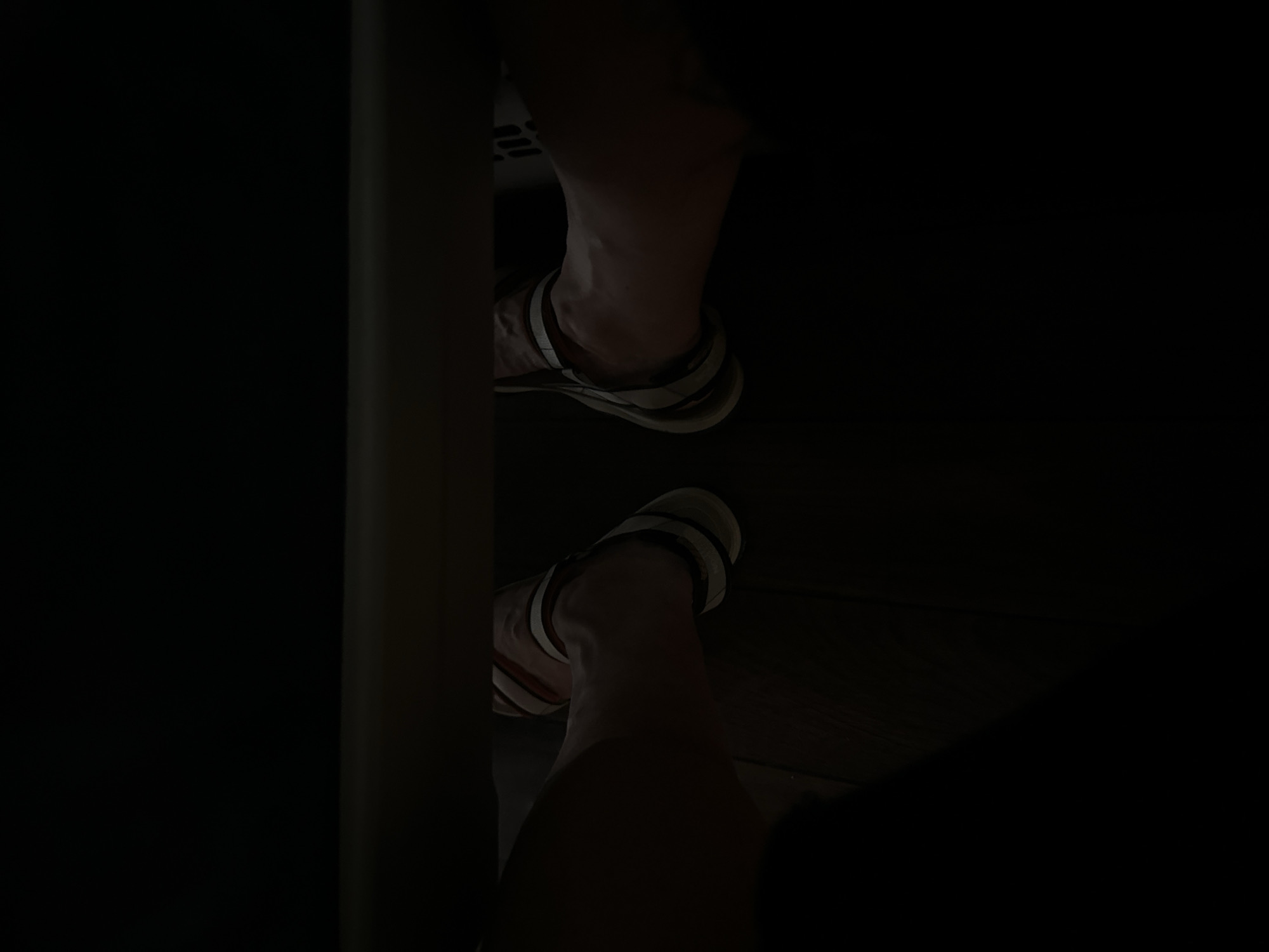 The image shows a close-up, low-angle view of a person's legs and feet, wearing sandals. The scene is dimly lit, with a horizontal dark surface in the foreground, possibly a railing or table edge, obscuring the upper portion of the frame. The floor appears to be dark wood. The subject's feet are positioned apart, and their skin tone is visible in the limited light. The sandals have a multi-striped design. The overall setting is dark, with some light illuminating the subject's lower legs and feet. No identifiable landmarks or context to pinpoint a location in Barcelona, Spain are present in the image.