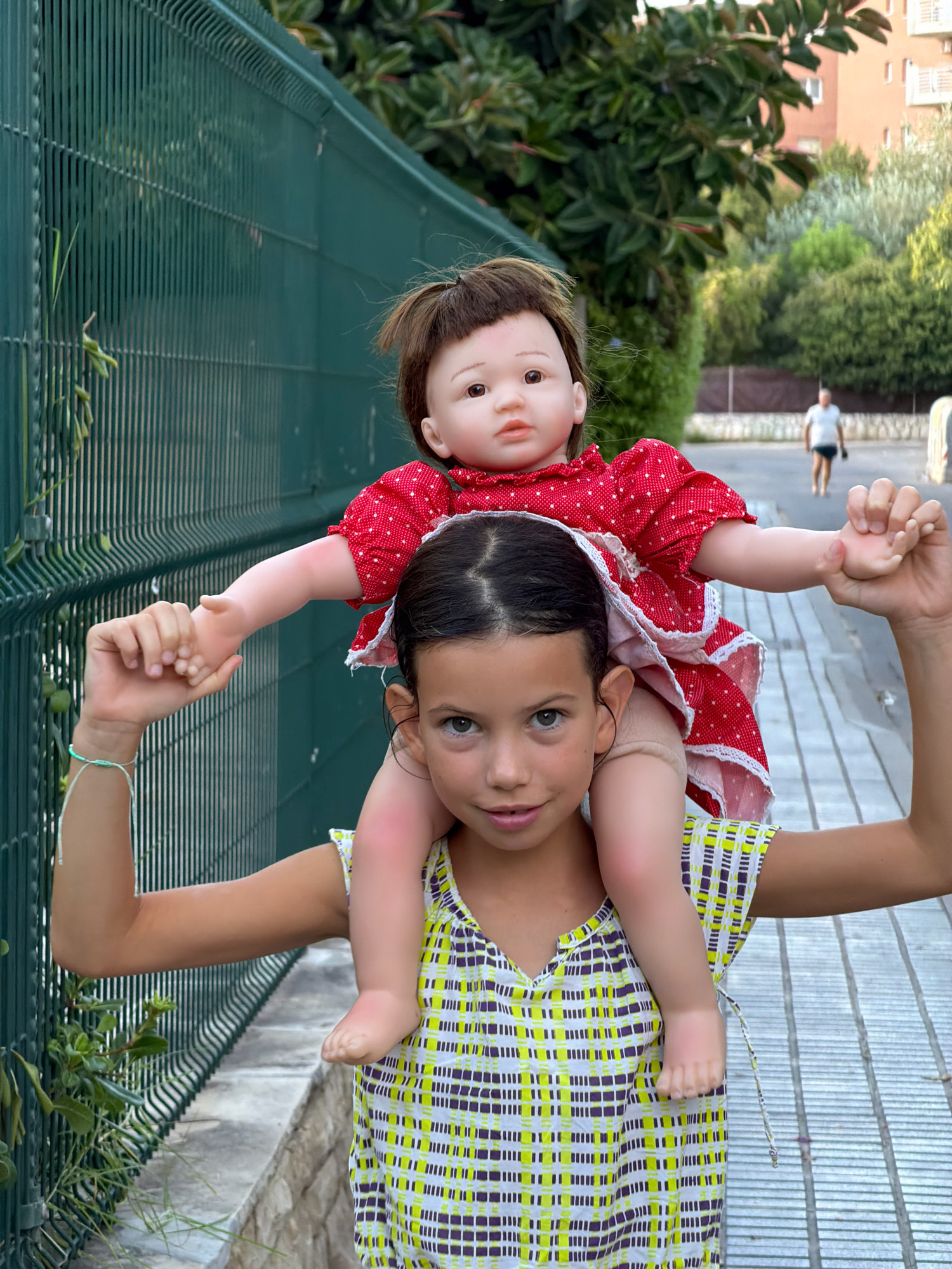 A young girl carries a life-sized doll on her shoulders outdoors in Salou, Spain. The girl, with dark hair pulled back and fair skin, is wearing a sleeveless, V-neck dress with a yellow, white, and purple checkered pattern. She is looking towards the camera with a slight smile. The doll, dressed in a red and white polka dot dress with lace trim, has brown hair and is positioned on the girl's shoulders. The doll's arms are outstretched, with one hand holding the girl's hand.
The background features a green metal fence on the left and a sidewalk with a gray, stone-like border on the right. Beyond the fence, lush green foliage and trees are visible, along with a glimpse of a residential building with balconies. Further in the distance, a person in a white t-shirt and dark shorts is walking away from the camera. The lighting suggests it is daytime, likely late afternoon given the soft light and shadows.