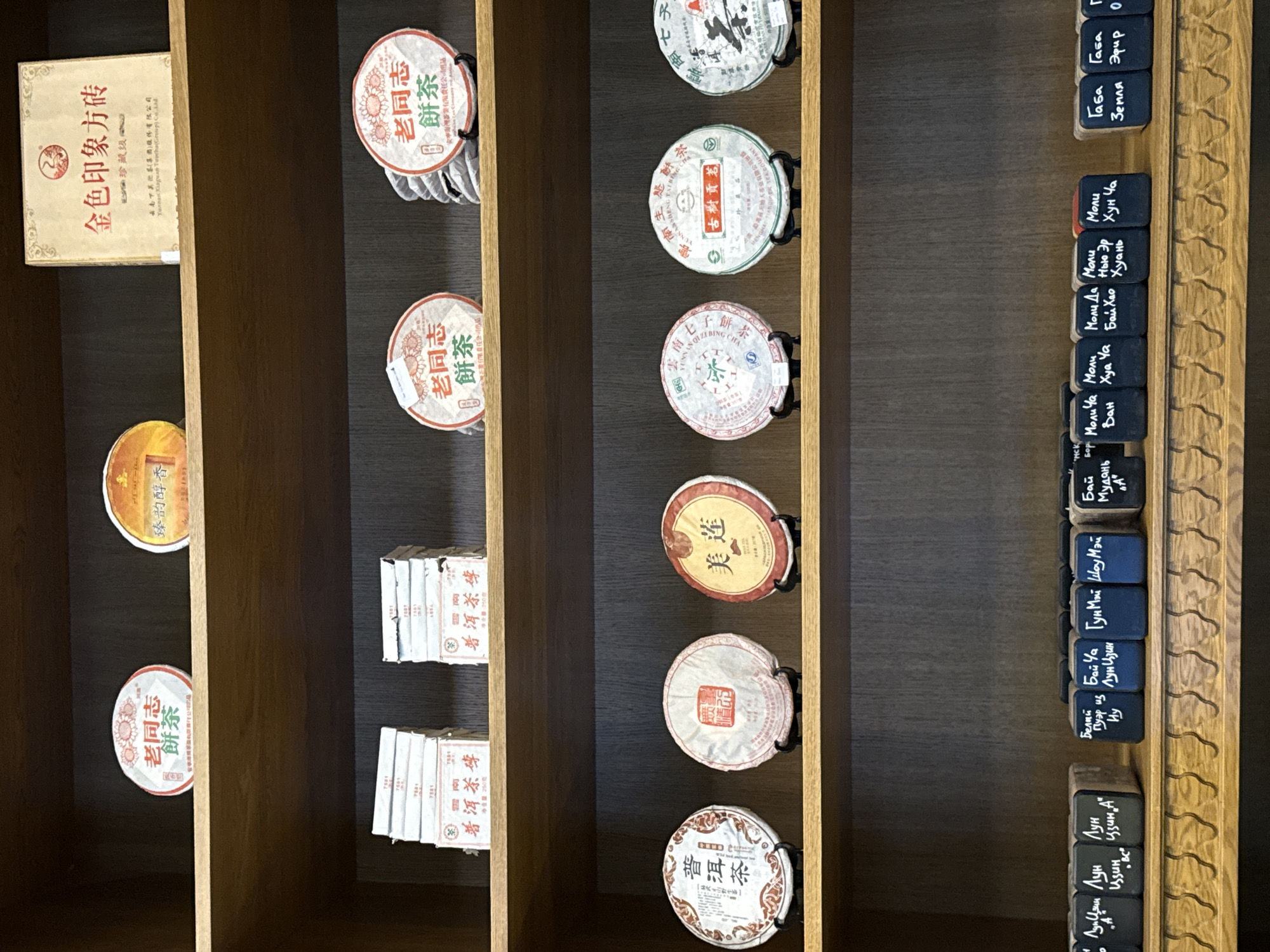 An interior shot captures a well-organized display of various Chinese teas on dark wooden shelves, likely within a tea shop in Moscow, Russia. No people are visible.
The shelves are filled predominantly with compressed tea cakes, primarily Puerh tea, identified by their round shape and traditional Chinese packaging. These cakes, varying in color and design, are either stacked or held upright in individual stands. Prominent labels on the tea cakes include "老同志 餅茶" (Old Comrades Cake Tea), "云南七子饼茶" (Yunnan Seven-Son Cake Tea), "古樹貢茶" (Ancient Tree Tribute Tea), and "普洱茶" (Puerh Tea). One cream-colored box is labeled "金色印象方砖" (Golden Impression Square Brick).
Beneath some of the round tea cakes, smaller, individually wrapped tea bricks are stacked. Their packaging reads "普洱茶磚 净含量:250克" (Puerh Tea Brick Net Weight: 250 grams).
On the far right, a vertical section of the display features numerous small, dark blocks, each with handwritten white Cyrillic text, likely indicating different tea types. These labels include "Молича Мудань A" (Jasmine Tea Peony A), "Моли Хуа Ча" (Jasmine Flower Tea), "Моли Бай Хао" (Jasmine White Hair), "Лунь Цзин А" (Longjing A), "Байча" (White Tea), "Гун Мэй" (Gongmei), "Шоу Мэй" (Shoumei), "Бай Мудань" (White Peony), "Пуэр" (Puerh), "Габа Земля" (GABA Earth), and "Габа Эфир" (GABA Ether). This combination of Chinese characters and Cyrillic script strongly suggests the shop caters to a Russian-speaking clientele interested in authentic Chinese teas.
The lighting appears to be artificial, evenly illuminating the products. The overall scene conveys a sense of a specialized and curated tea collection.