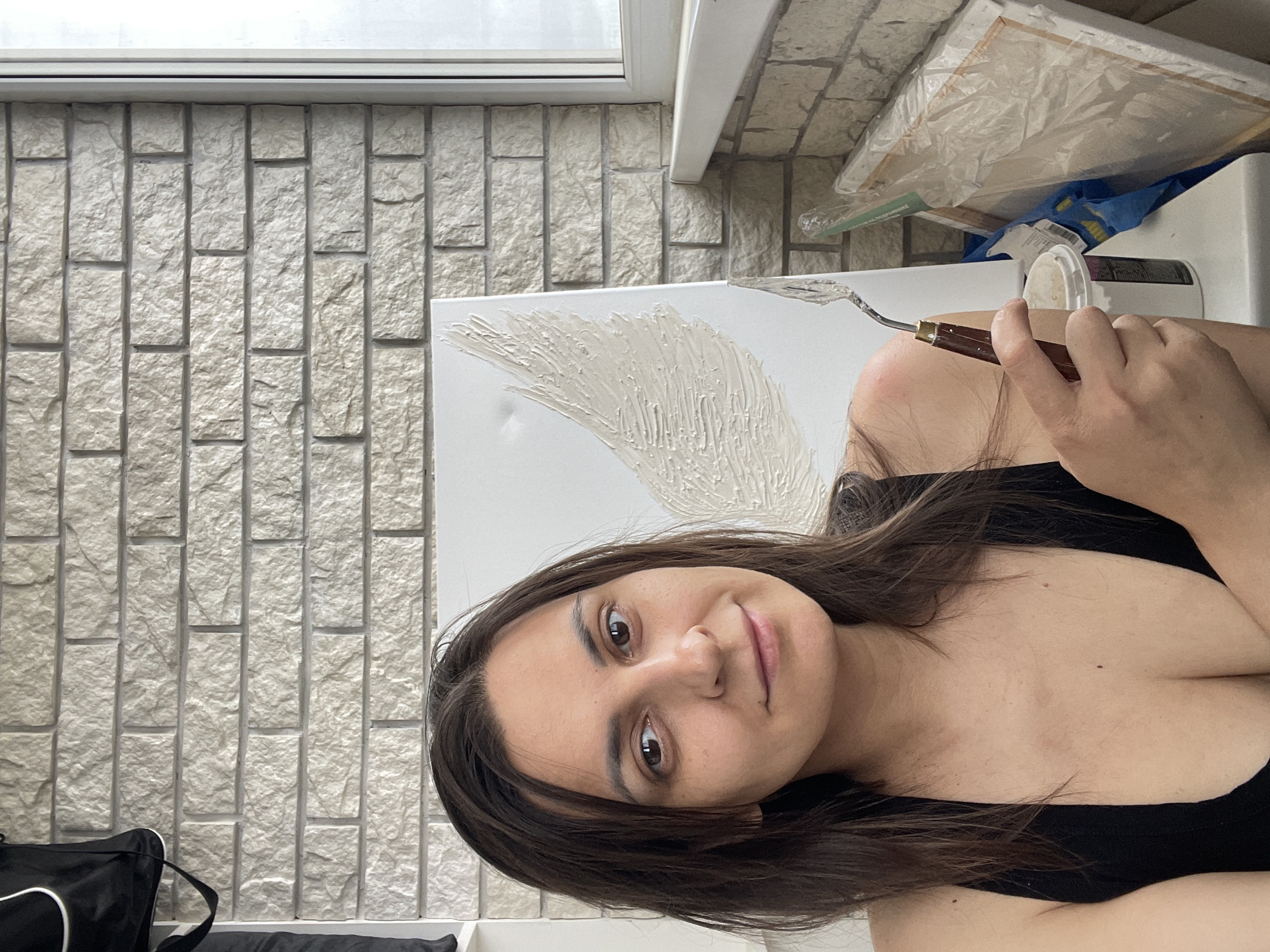 A portrait of a woman holding a painting knife in front of a textured painting. The woman has long brown hair and brown eyes and is wearing a black tank top. The painting appears to have raised white paint applied in an abstract design. Behind the woman and the painting is a wall with a brick-like pattern. Near the top of the frame is a windowsill and some art supplies including canvas frames wrapped in plastic. The location is Unknown, Unknown.