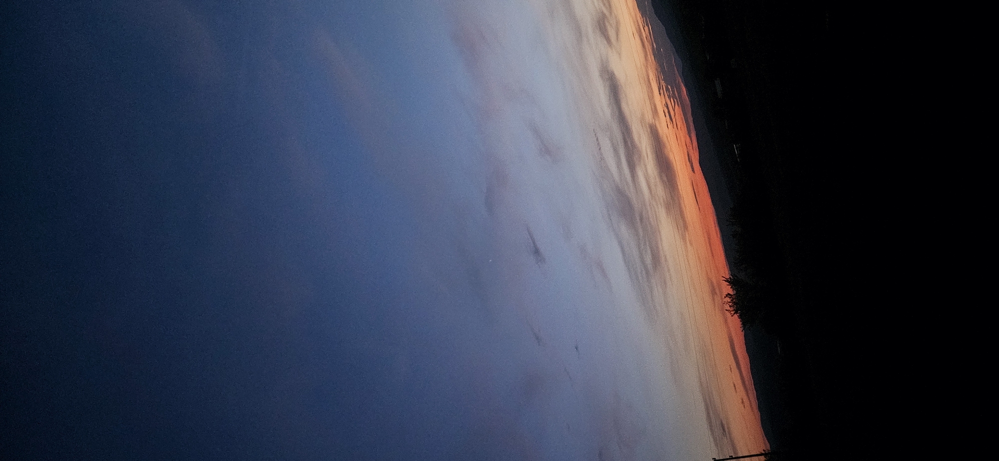 The image depicts a sunset over a rural landscape, likely Momin Prohod, Bulgaria. The sky transitions from a deep navy blue at the top to lighter blues and then streaks of orange and pink near the horizon. Thin, wispy clouds are scattered across the sky. Silhouetted against the sunset is a dark, undulating treeline and hills. In the foreground, a dark, silhouetted object, possibly a utility pole, is visible on the left side. The bottom portion of the image is entirely black, obscuring any ground-level details. There are no people or visible actions taking place.