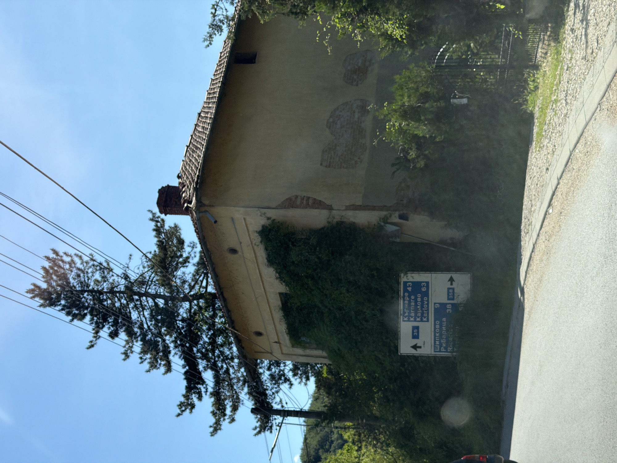 This image depicts a view of a street with a building and surrounding vegetation in Beli Osam, Bulgaria. The building is a two-story structure with a light yellow facade and a tiled roof. The right side of the building shows exposed brickwork where the plaster has fallen off. The building is partially overgrown with dense green ivy. In front of the building, there are two directional road signs. The upper sign indicates "Kurnare 43", "Karnare", "Karlovo 63" with an upward arrow and the number 35. The lower sign indicates "Shipkovo 9", "Ribaritsa" with an arrow pointing to the right and the numbers 38 and 356. A tall evergreen tree stands to the left of the building, and power lines crisscross the sky above. To the right of the building, there is a black metal gate with a paved walkway leading to it. The scene is set under a clear blue sky with sunlight casting shadows. The foreground shows a paved road with a curb.