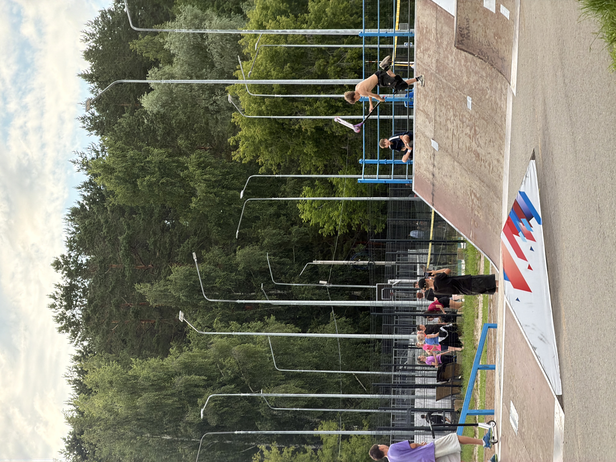 The image depicts an outdoor scene at a skatepark, possibly in Chaikovsky, Russia, as suggested by the location context. The central focus is a ramp structure, likely made of wood and concrete, with a red and white banner featuring red, blue, and white geometric shapes attached to its side.
Several people are visible, engaged in or spectating activities at the skatepark. In the foreground, a young person in a purple shirt and gray shorts stands with a scooter. Further up the ramp, a shirtless young person with blond hair is in mid-air on a scooter, performing a trick. Another individual, a young person in a dark shirt and shorts, is crouched on the ramp. Behind the ramp, a group of people, including individuals wearing colorful tops like pink and blue, are gathered, observing the events. A person in a black shirt and shorts is standing near the banner, possibly taking a photo or observing.
The setting is characterized by a clear sky with scattered clouds, suggesting daytime, likely late afternoon or early evening given the lighting. Lush green trees form a dense backdrop, indicating a park or natural setting adjacent to the skatepark. Tall streetlights are also a prominent feature in the background, lining the area. The overall atmosphere appears to be casual and active, with a focus on extreme sports. No visible text other than that on the banner is legible.