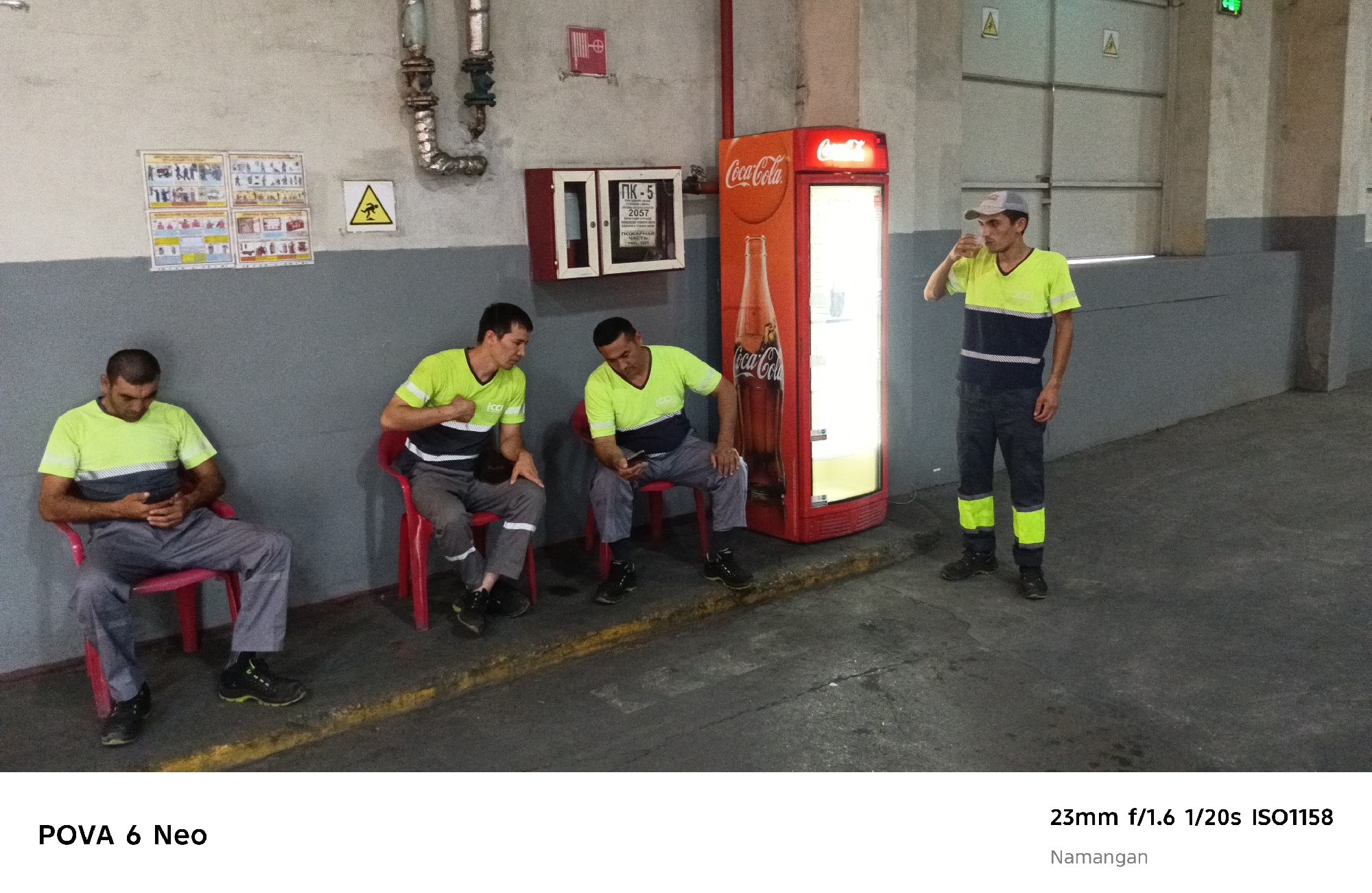 In Namangan City, Uzbekistan, four men in work uniforms are in an indoor industrial or storage area. Three men are seated on red chairs, and one is standing. The seated men are looking down at their phones or resting. The standing man is drinking from a cup, wearing a baseball cap and a fluorescent yellow and navy blue shirt with reflective stripes on the pants. A red Coca-Cola vending machine is to his right. The environment features concrete walls, a concrete floor, and some industrial piping on the wall. There are safety signs on the wall above the seated men.