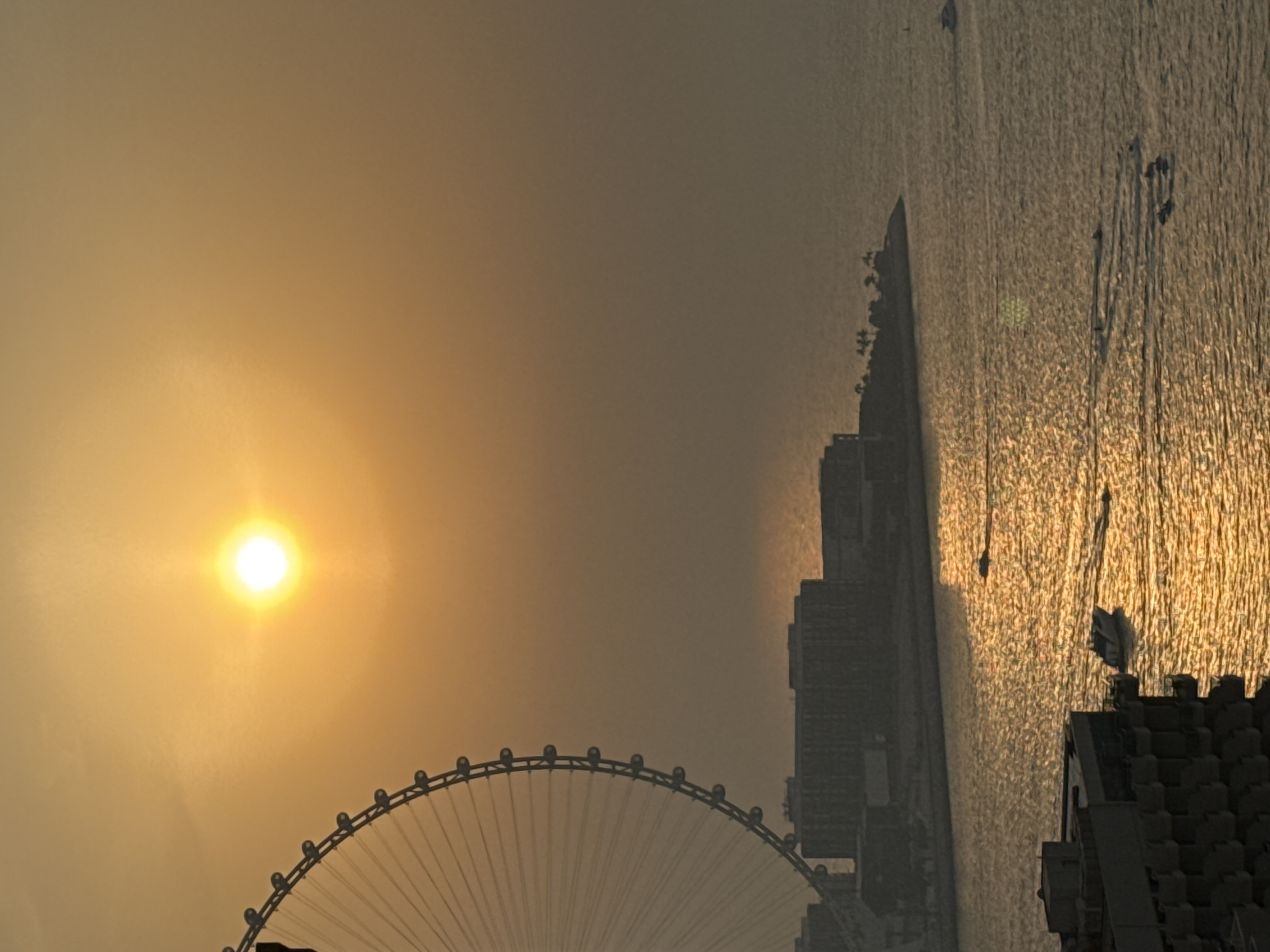 This image captures a sunset over the waters of Dubai, United Arab Emirates, likely from the JBR (Jumeirah Beach Residence) area. A large Ferris wheel, possibly Ain Dubai, is silhouetted on the left side of the frame, its spokes extending outwards. In the middle ground, a cluster of modern buildings, characteristic of Dubai's skyline, is visible. The setting sun casts a warm, golden glow across the water, creating a shimmering effect. Several boats, including a jet ski formation and individual speedboats, are seen traversing the water, leaving wakes behind them. The overall atmosphere is one of a vibrant urban waterfront at dusk.