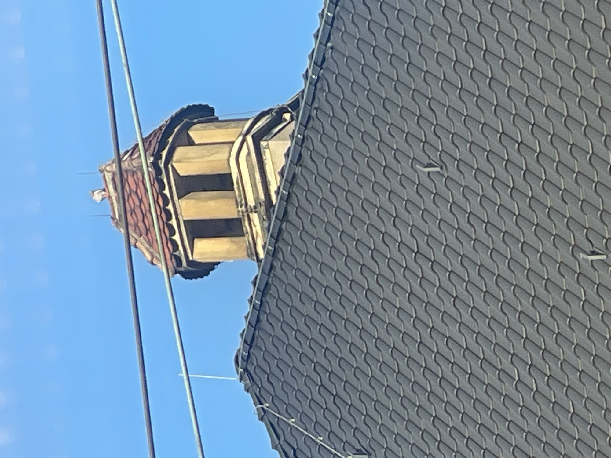 A close-up shot captures a section of a building's rooftop against a clear blue sky. The foreground is dominated by a dark grey, tiled roof, with the tiles arranged in a uniform, overlapping pattern. Several metal elements, likely part of a lightning protection system, protrude from the roof.
Further up, a distinctive architectural feature stands out. It appears to be a small, octagonal tower or turret with a red-tiled, conical roof. The tower has multiple openings or windows, each framed by light-colored, possibly stone or stucco, trim. A small, light-colored object, possibly a bird or a decorative finial, is perched on the very top of the turret.
Two parallel horizontal wires, dark in color, stretch across the upper portion of the frame, obscuring a small part of the sky and the top of the turret. The overall scene suggests an urban setting, possibly in Hagen, Germany, given the architectural style of the turret and the presence of overhead wires often seen in European cities. The lighting indicates it is daytime, with sunlight casting subtle shadows on the roof tiles.