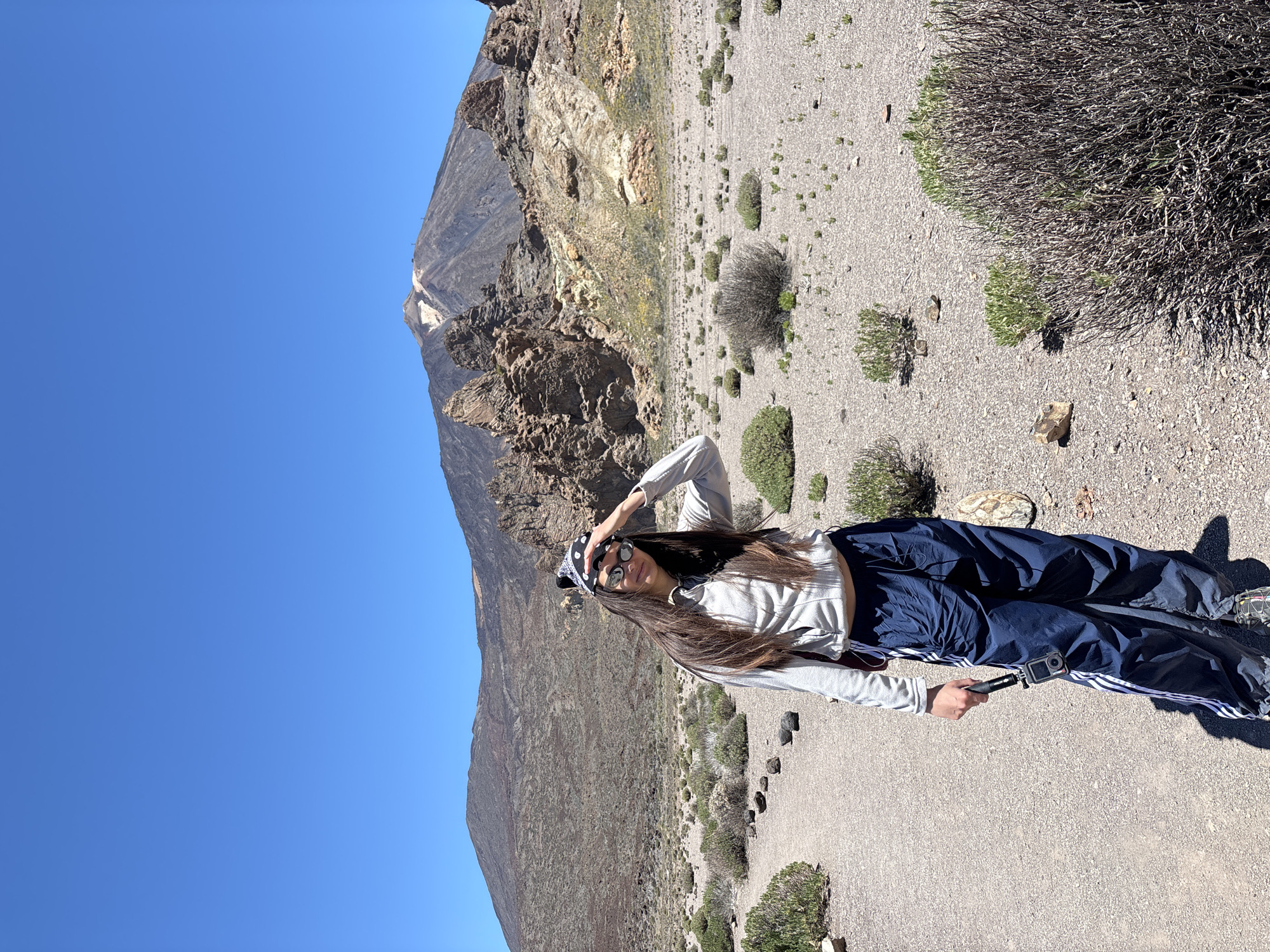A woman stands on a gravelly path in a volcanic landscape, possibly in Teide National Park, La Orotava, Spain. She wears sunglasses and a bandana, a light gray long-sleeved shirt, and blue track pants with white stripes. She holds a camera on a selfie stick in her right hand and raises her left hand to shield her eyes from the sun. Behind her, a large, rocky mountain dominates the view, set against a clear blue sky. The surrounding terrain is sparsely vegetated with low shrubs.