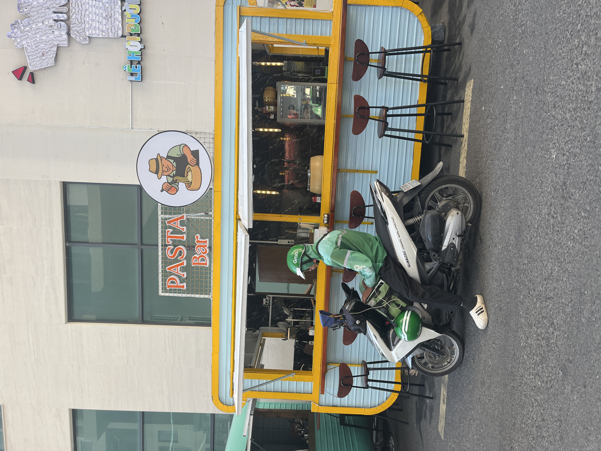 A food vendor operating a mobile "Pasta Bar" on a street in Bac Nha Trang Ward, Vietnam, is interacting with a Grab delivery driver on a white scooter. The driver, clad in a green Grab uniform and helmet, is positioned on the vehicle near the bar, where several stools are placed. The Pasta Bar, painted with a yellow and light blue exterior, features a window with a food illustration and the text "Pasta Bar." The background includes a multi-story building and glimpses of nearby objects. The street appears to be paved and the scooter is parked parallel to the curb.