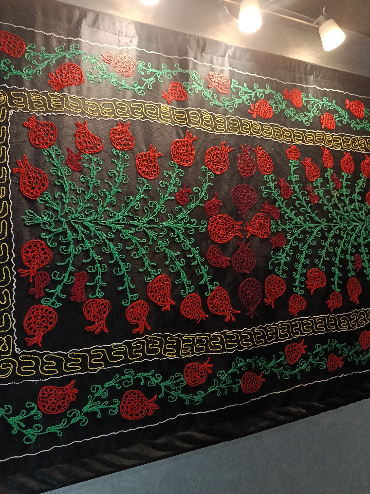 A close-up shot reveals a decorative fabric hanging on a wall. The fabric is predominantly black, embellished with a pattern of embroidered pomegranates in shades of red and burgundy, interspersed with green vines. A band of decorative stitching in yellow and white runs horizontally across the fabric. The fabric is possibly part of an exhibition in the city of Moscow, Russia, but further information is not included.