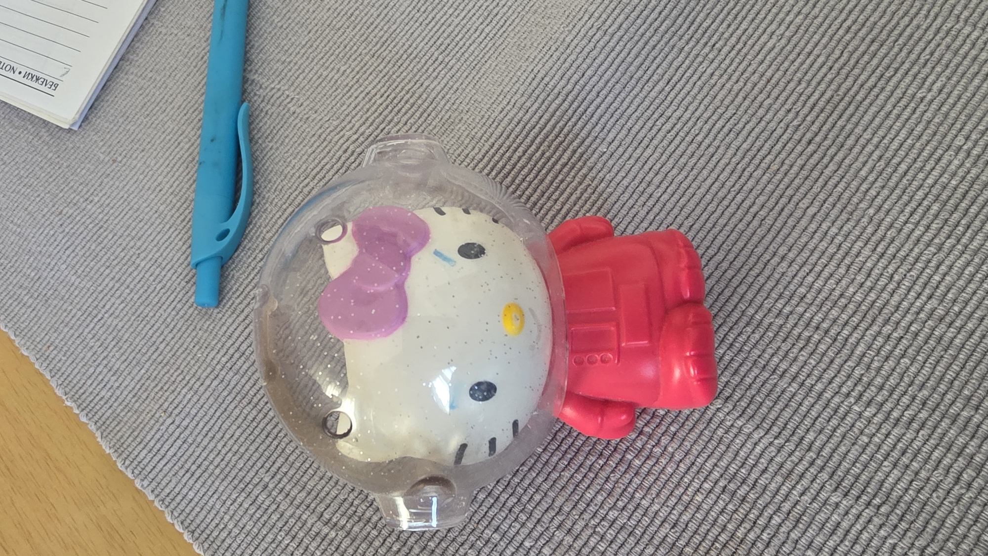 The scene is an indoor setting, likely a table, covered with a gray fabric with a woven texture. In the center of the frame is a Hello Kitty toy inside a clear plastic sphere attached to a red plastic piece. The toy appears to be wearing a space helmet. A blue pen is partially visible to the left of the toy, and a corner of a notepad is visible in the upper left corner. The description of the location is not provided in the prompt and can therefore not be provided.