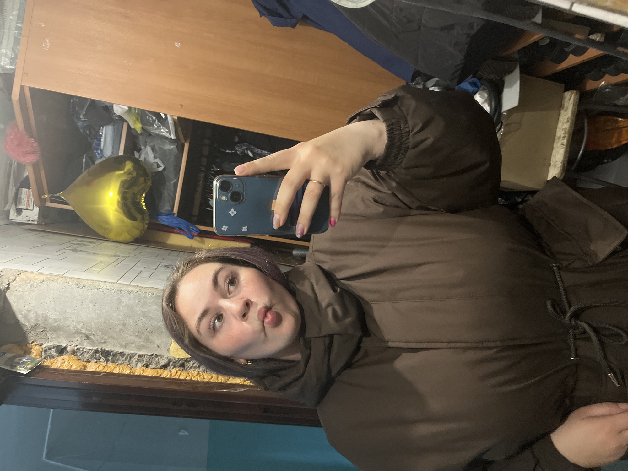 A woman with brown hair is taking a selfie with a blue iPhone in a cluttered room. She is wearing a brown puffy jacket and making a kissing face. Behind her, there's a wooden cabinet filled with various items, including a gold heart-shaped balloon. The room appears to be poorly lit and under renovation with exposed construction material. The location is likely in the city of Chaikovsky, Russia.