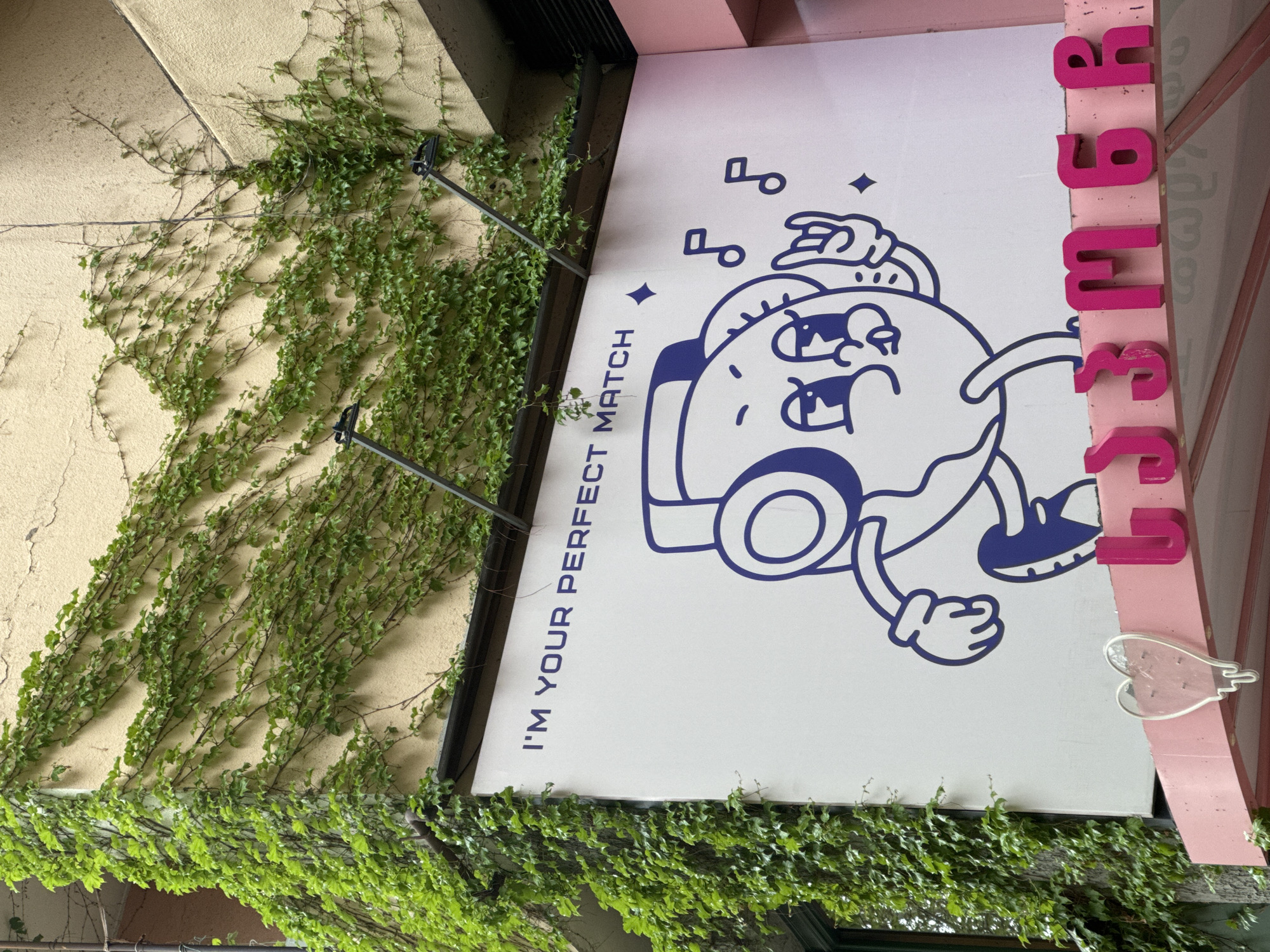 A close-up, slightly angled shot depicts a sign attached to a building exterior, partially obscured by green vines. The sign features a cartoon-like image of a donut with anthropomorphic features - eyes, mouth, arms, and legs - drawn in blue on a white background. Above the donut are musical notes and stars. The text on the sign reads "I'M YOUR PERFECT MATCH" in capital letters. To the right of the sign, affixed to a pink-painted structure, are vertically arranged pink letters spelling "333 M6A." A strawberry shaped sign with a heart shaped hole cut out is underneath the text on the sign. The building behind the sign appears to be light yellow with a rough texture. The location is Tbilisi, Georgia.