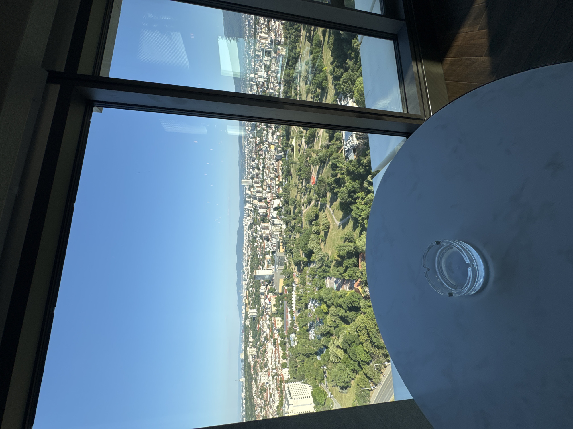 The image shows a view from a high-rise building overlooking the city of Sofia, Bulgaria. In the foreground, a dark, round table is visible with an empty glass ashtray placed on its surface. The table is positioned near a large window that offers a panoramic view of the cityscape. The city appears densely populated with a mix of buildings, ranging from smaller residential structures to taller modern office buildings. A large green park area is prominent in the middle ground, with trees and pathways. Beyond the city, a range of mountains can be seen in the distance under a clear blue sky. The overall scene suggests a clear, sunny day.