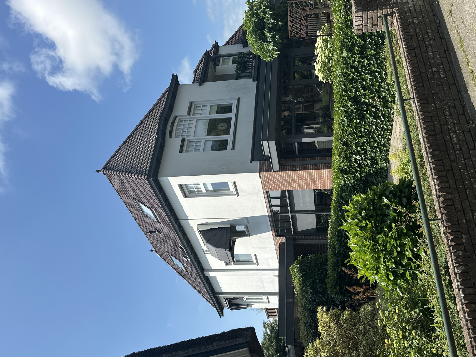 A two-story white stucco house with a dark tiled roof and dormer windows is situated in a suburban environment under a clear blue sky with scattered white clouds. The house features multiple windows, including a large bay window on the second story with a decorative transom. A dark retractable awning is extended over a ground-floor window. The ground level of the house incorporates a section with red brickwork and large glass windows, suggesting a conservatory or extension. Lush green vegetation, including hedges and shrubs, surrounds the property, with a brick wall and a green metal railing in the foreground. The scene is likely captured on a sunny day.