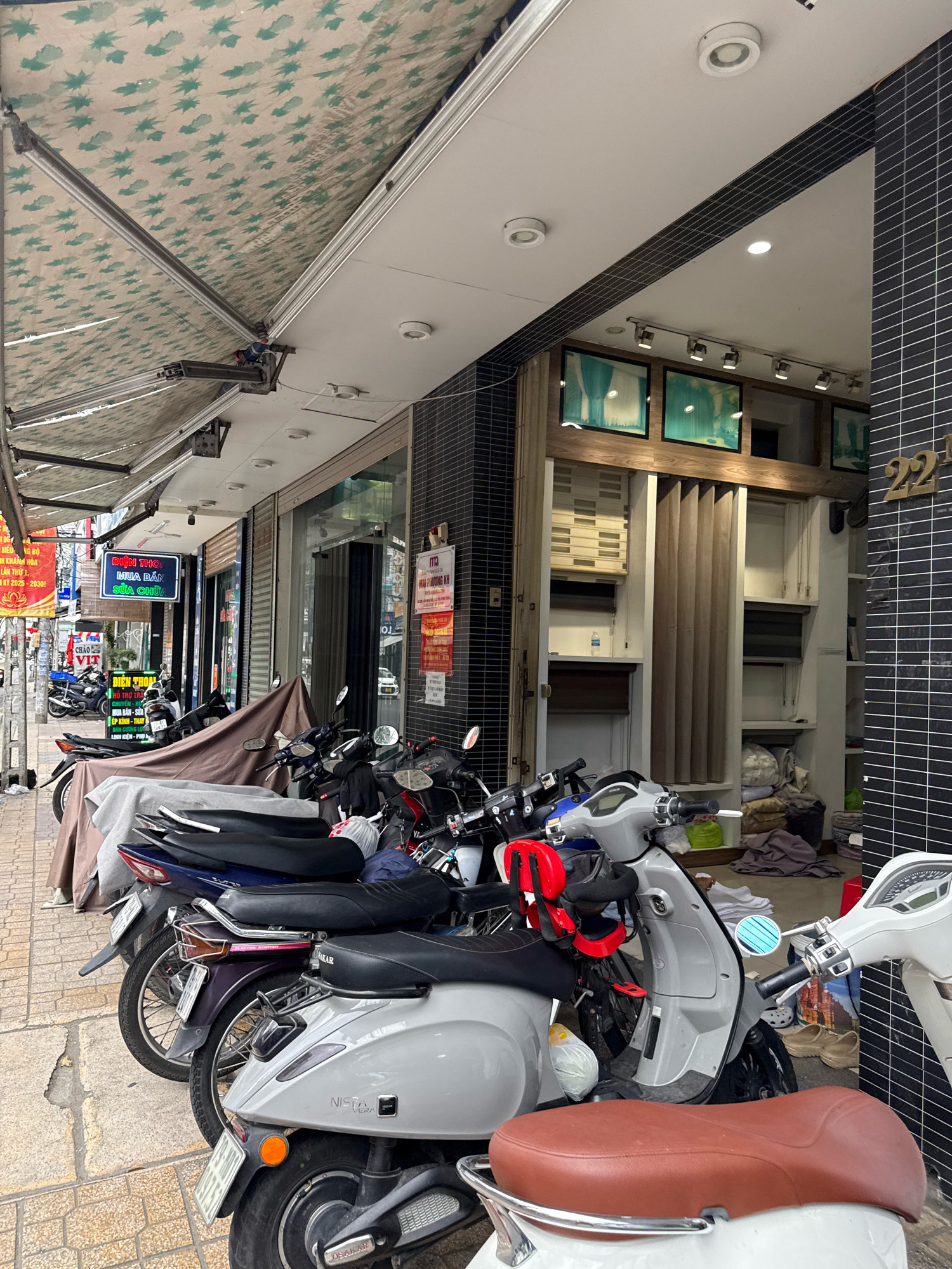 This is a medium shot of a row of parked motorcycles in front of a shop on a sidewalk in Nha Trang, Vietnam. The motorcycles are lined up along the building, some covered with tarps. The shop has a black tiled facade with a sign that has the number 22 on it. There are framed pictures and blinds visible inside the shop. The sidewalk is made of light-colored square pavers. An awning with a green and white leaf pattern extends over the sidewalk from the building. There are no people visible in the image.