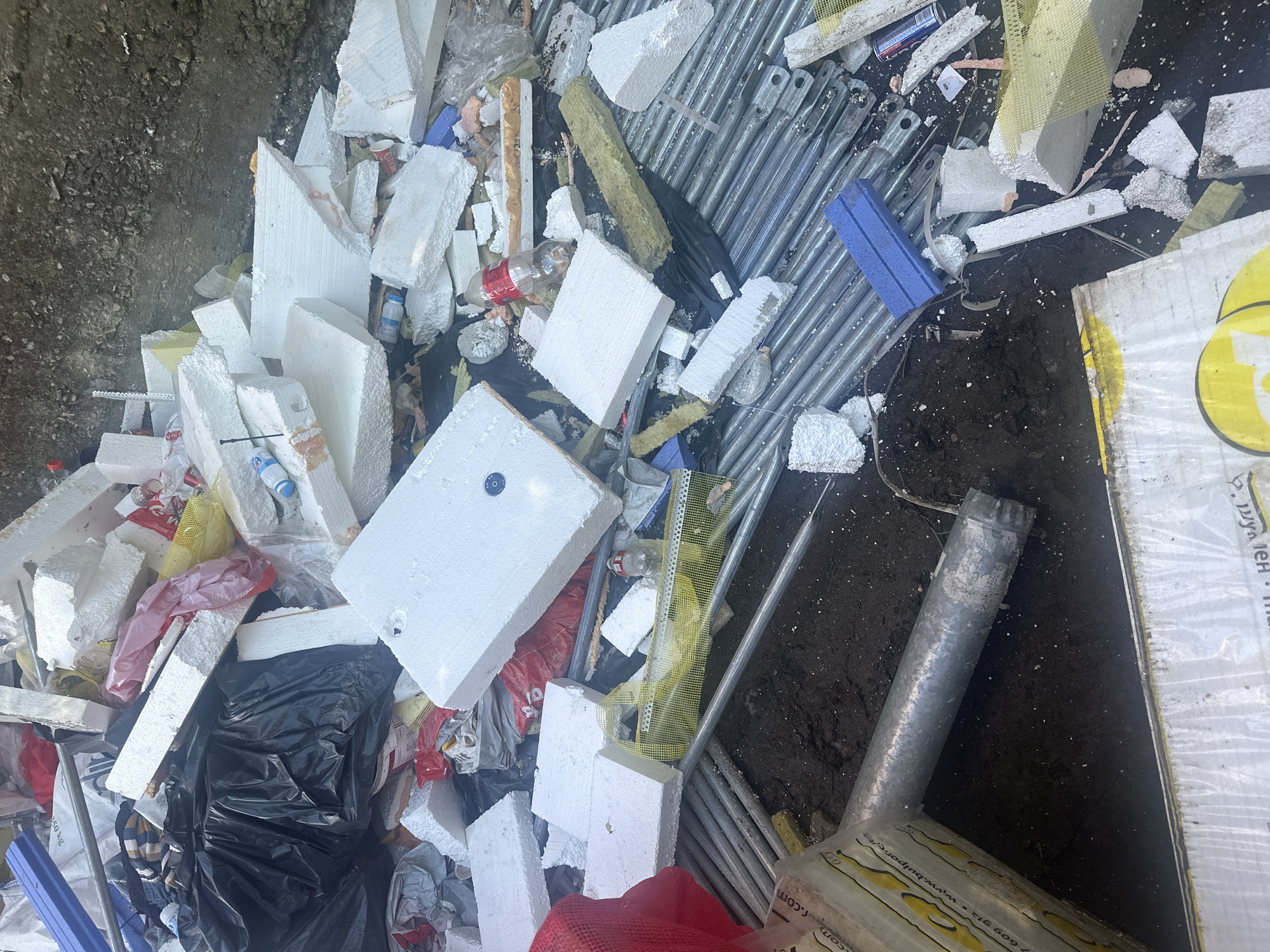 A pile of construction debris sits on the ground. The debris includes large pieces of white styrofoam, black trash bags, metal poles, yellow mesh, and various other items. The ground appears to be dirt and gravel. The scene is outdoors, and the lighting suggests it is daytime.