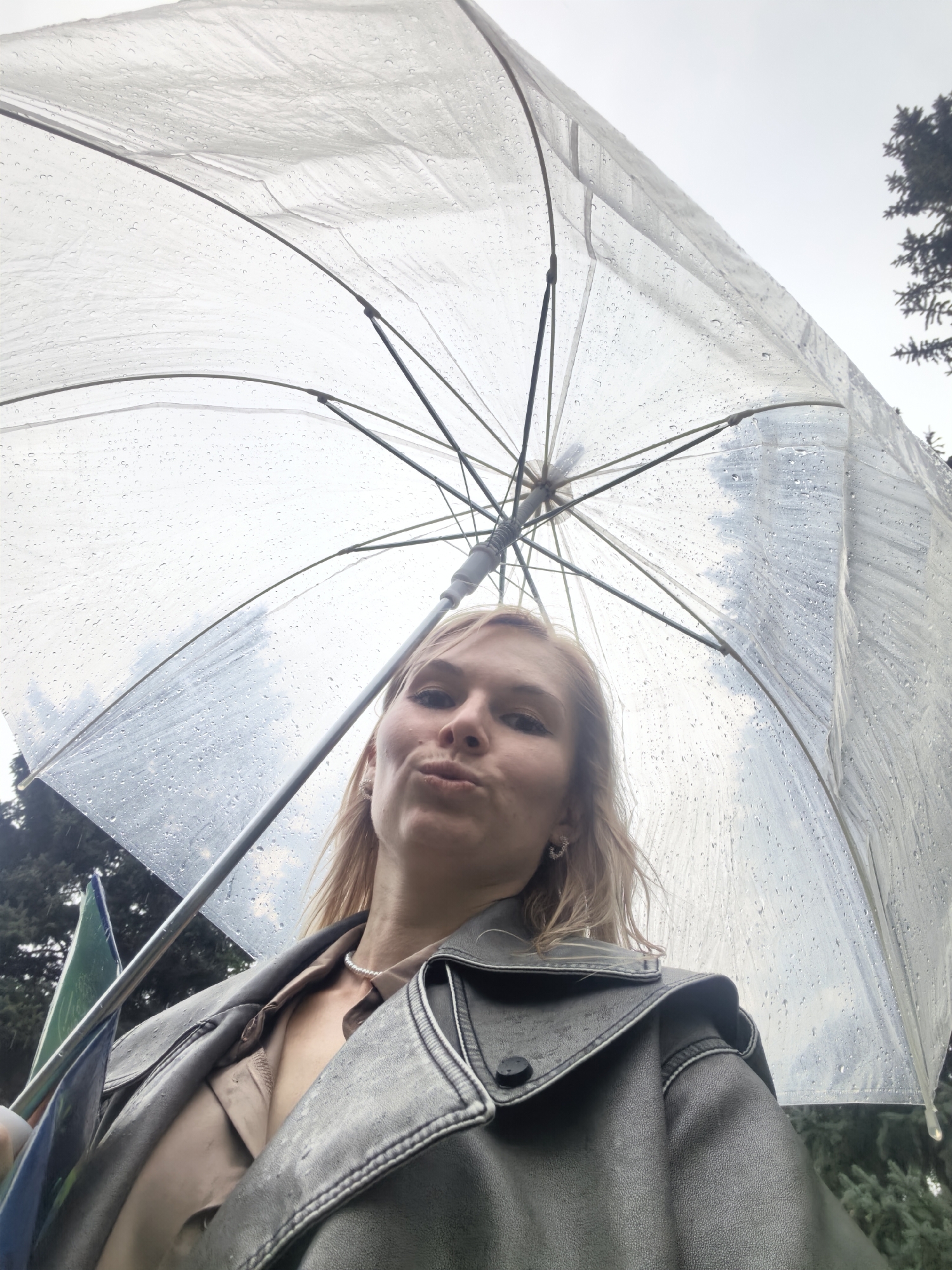 A young woman with blonde hair is taking a selfie under a clear umbrella. She is wearing a beige collared shirt and a dark, possibly leather, jacket. The umbrella is wet with raindrops. In the background, trees are visible, suggesting an outdoor setting. The scene takes place in Sterlitamak, Russia.