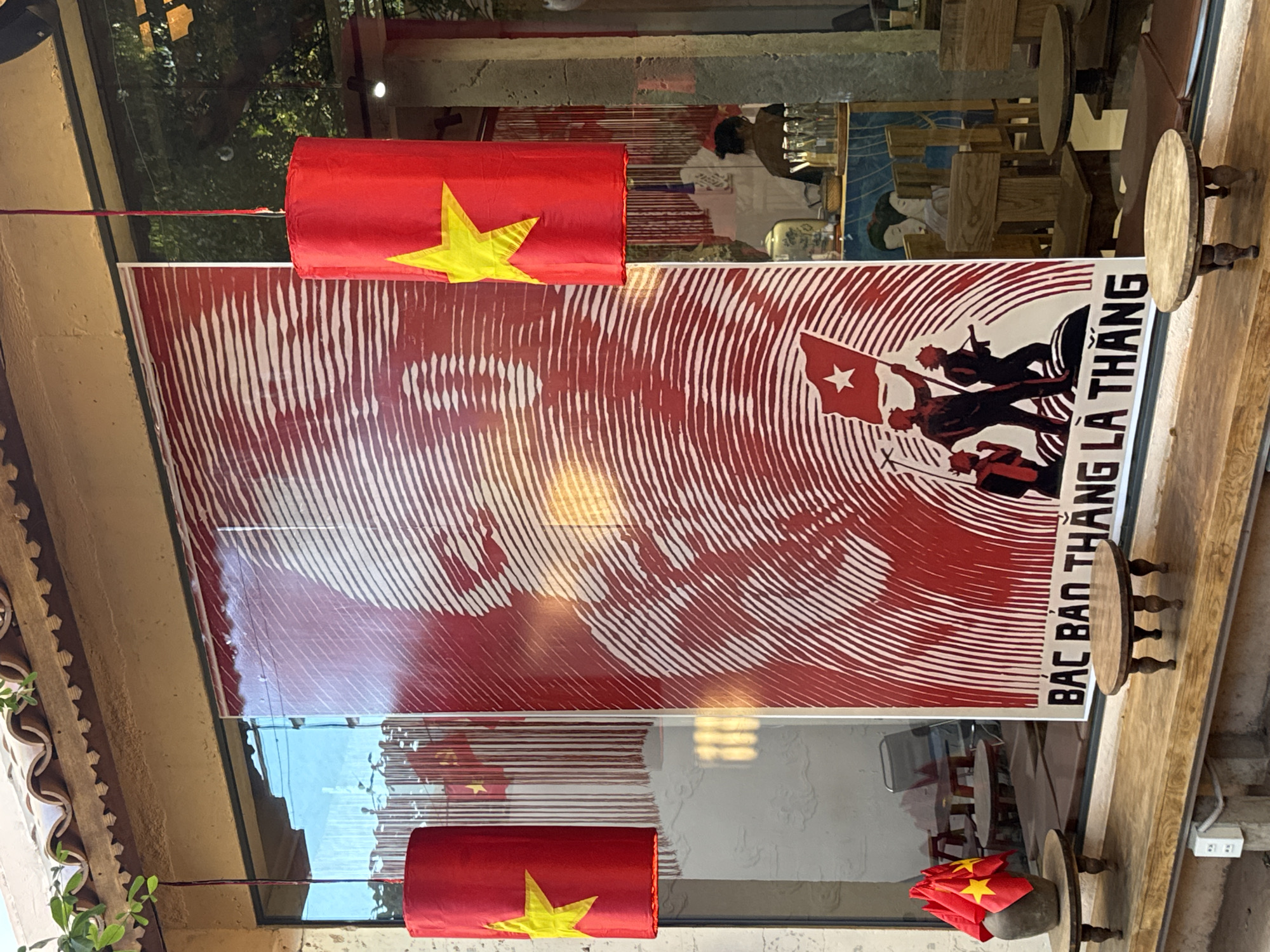 The image displays the exterior facade of a commercial establishment, likely a cafe or shop, located in Nha Trang, Vietnam. A large, vertical red and white poster occupies the central portion of a wide glass window. The poster features a stylized, line-art portrait of Ho Chi Minh in its upper two-thirds, rendered with white horizontal wavy lines on a red background. The lower third depicts three silhouetted figures of soldiers on a white and red striped ground, with the central figure holding a red flag adorned with a white star. White text "BÁC BẢO THẮNG LÀ THẮNG" is printed horizontally at the bottom of the poster.
Two red cylindrical lanterns, each displaying a yellow five-pointed star, hang by red cords from the building's overhang, positioned to the left and right of the main poster. Below the window, a long, light-colored wooden bench or shelf extends across the facade. On this bench, to the left, a dark, rustic-looking pot holds several small Vietnamese flags. Two small, round, low wooden tables with four carved legs are also placed on this bench. A white electrical outlet is mounted on the wall beneath the bench on the far left.
Through the clear portions and reflections on the glass window, the interior of the establishment is partially visible. Wooden chairs and tables are discernible. On the right side, a person with dark hair tied back is seen from the back, positioned behind what appears to be a counter or service area, with various bottles and containers present. The building features a light-brown tiled roof with a decorative, jagged-edged trim and some green foliage draped over it.