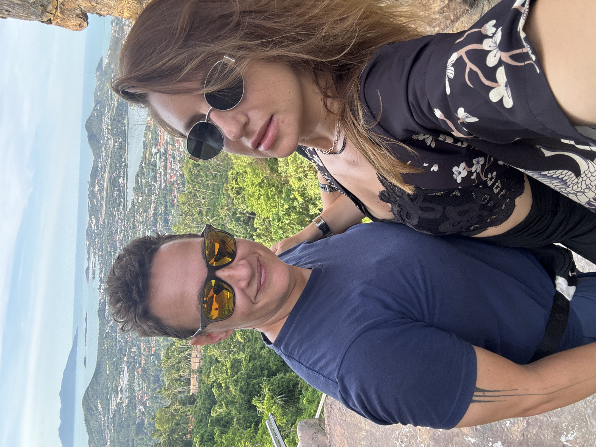 Two individuals, a person with short, dark hair wearing sunglasses and a navy blue t-shirt, and a person with long, brown hair wearing sunglasses and a black floral-patterned kimono over a black lace top, are taking a selfie. They are standing outdoors with a scenic view of a coastal city and islands in the background. The person on the left has a tattoo on their left arm. They appear to be on a rocky, elevated vantage point overlooking the city. The sky is partly cloudy.