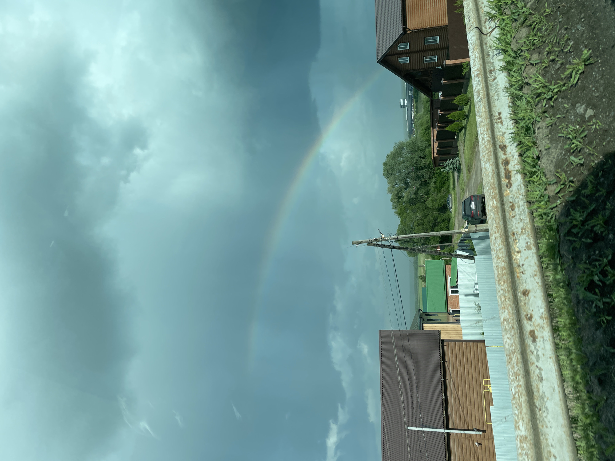 The image captures a rainbow arching across a cloudy sky in Malaya Ven'ya, Russia. A brown, multi-story house is on the right side of the frame, next to a road with a car parked. Other buildings with green and brown roofs can also be seen. A power line stretches across the image, and a rusty metal guardrail is in the foreground. The scene is photographed from an elevated perspective, facing slightly upward.