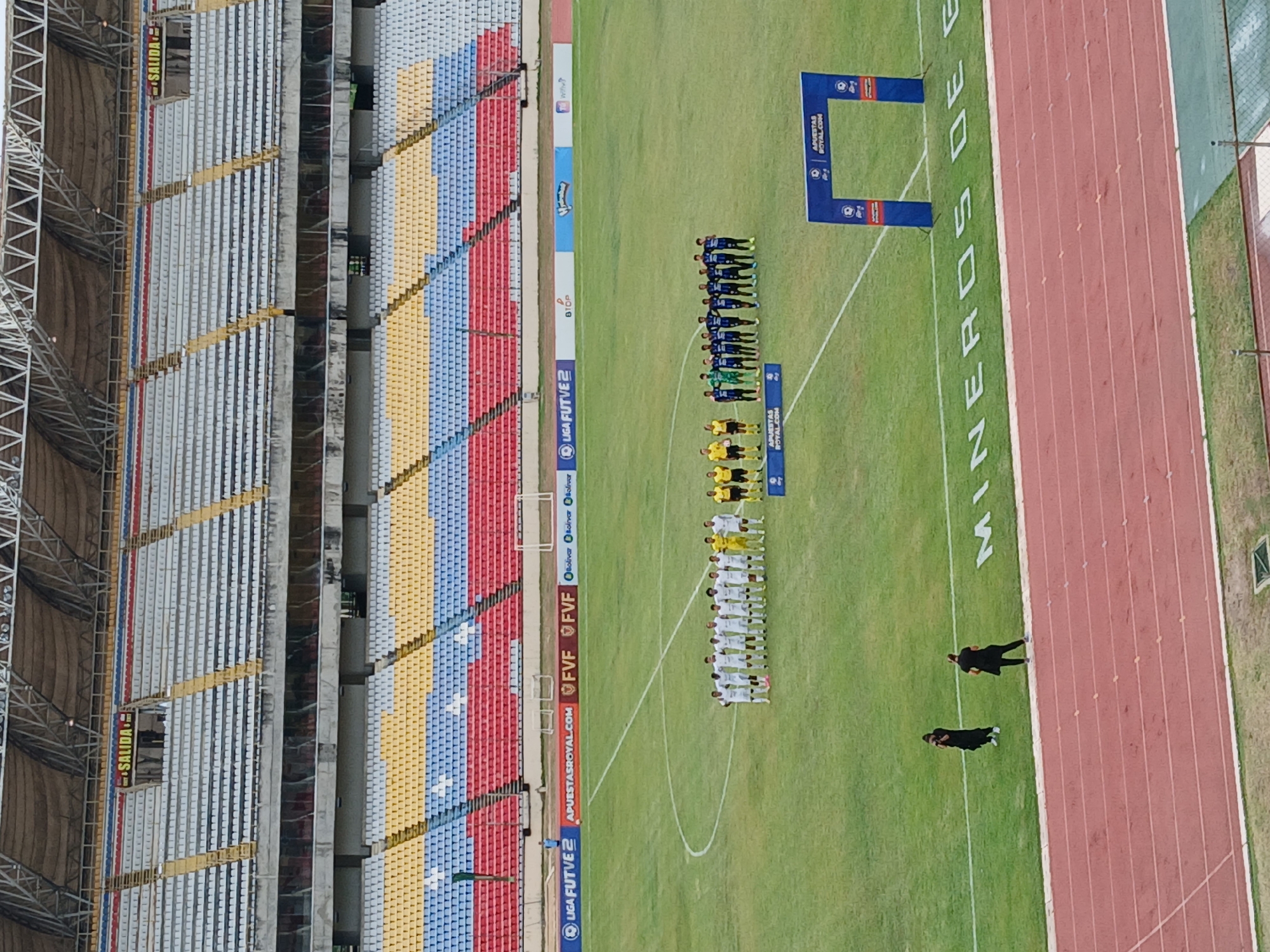 Here is a description of the image:
The image shows a soccer field and stadium. In the field's center, two teams of soccer players are lined up. One team is wearing white uniforms and the other is wearing blue. Behind the teams are two blue arched signs with the words "APUESTAS ROYAL.COM". To the right of the field is a red running track with the words "MINEROS DE" printed in large white letters on the green grass next to it. Two people dressed in black are walking on the field.
The stadium's seats are visible on the left. The seating is colored in red, blue, and yellow sections. A sign that says "SALIDA" is placed on a vertical column. Various sponsor logos and text are printed on the field's perimeter.