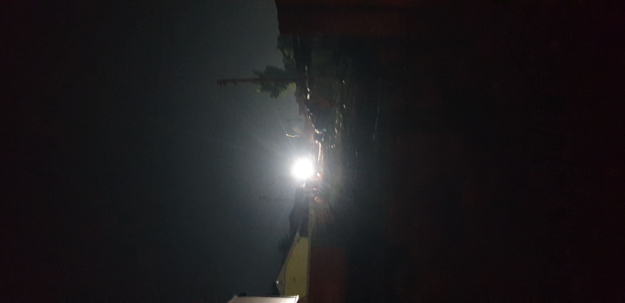 The image displays a night scene, oriented vertically with darkness dominating the left and right sides. When correctly viewed (rotated 90 degrees counter-clockwise), it presents an outdoor setting characterized by deep darkness punctuated by a single, extremely bright light source positioned slightly to the right of the center. This light emits a strong glare, illuminating a limited area immediately around it and creating visible light rays.
On the left side of the frame, silhouetted against the dark sky, are several trees, some resembling palm trees, and a tall, slender structure that appears to be a utility pole or communication mast. The upper portion of the scene is enveloped in darkness.
To the right of the central light, faint and indistinct outlines of buildings or structures are discernible. One structure in the lower right foreground features a visible segment of a yellowish wall. The area directly illuminated by the bright light suggests a ground or street surface, but details are obscured by the intense glow and overall low light conditions.
No specific people or activities are clearly visible within the scene, which primarily showcases the contrast between a powerful artificial light source and the surrounding darkness. The atmosphere immediately around the light appears slightly hazy. There is no visible text present in the image. The scene captures a moment of artificial illumination cutting through the profound darkness of night in what can be inferred as an outdoor environment in Sabon Tasha, Nigeria.
