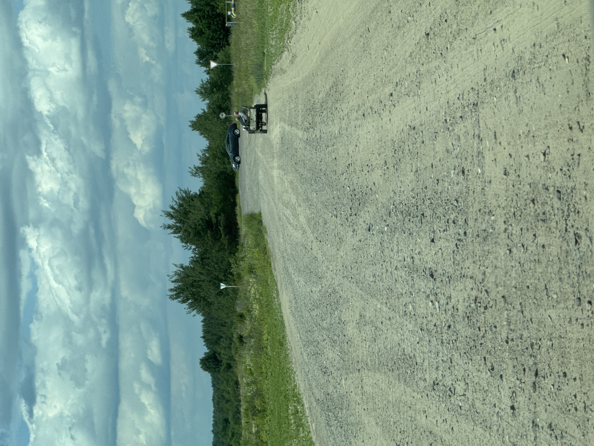 An outdoor, daytime, medium shot captures a gravel road leading into the distance, bordered by green grass and trees. A person is seated in a small, four-wheeled utility vehicle with a trailer, traveling on the road. In the background, a dark-colored car is parked on the side of the road. The sky is filled with dramatic, cloudy formations. The scene appears to be located in Orlivka, Ukraine.