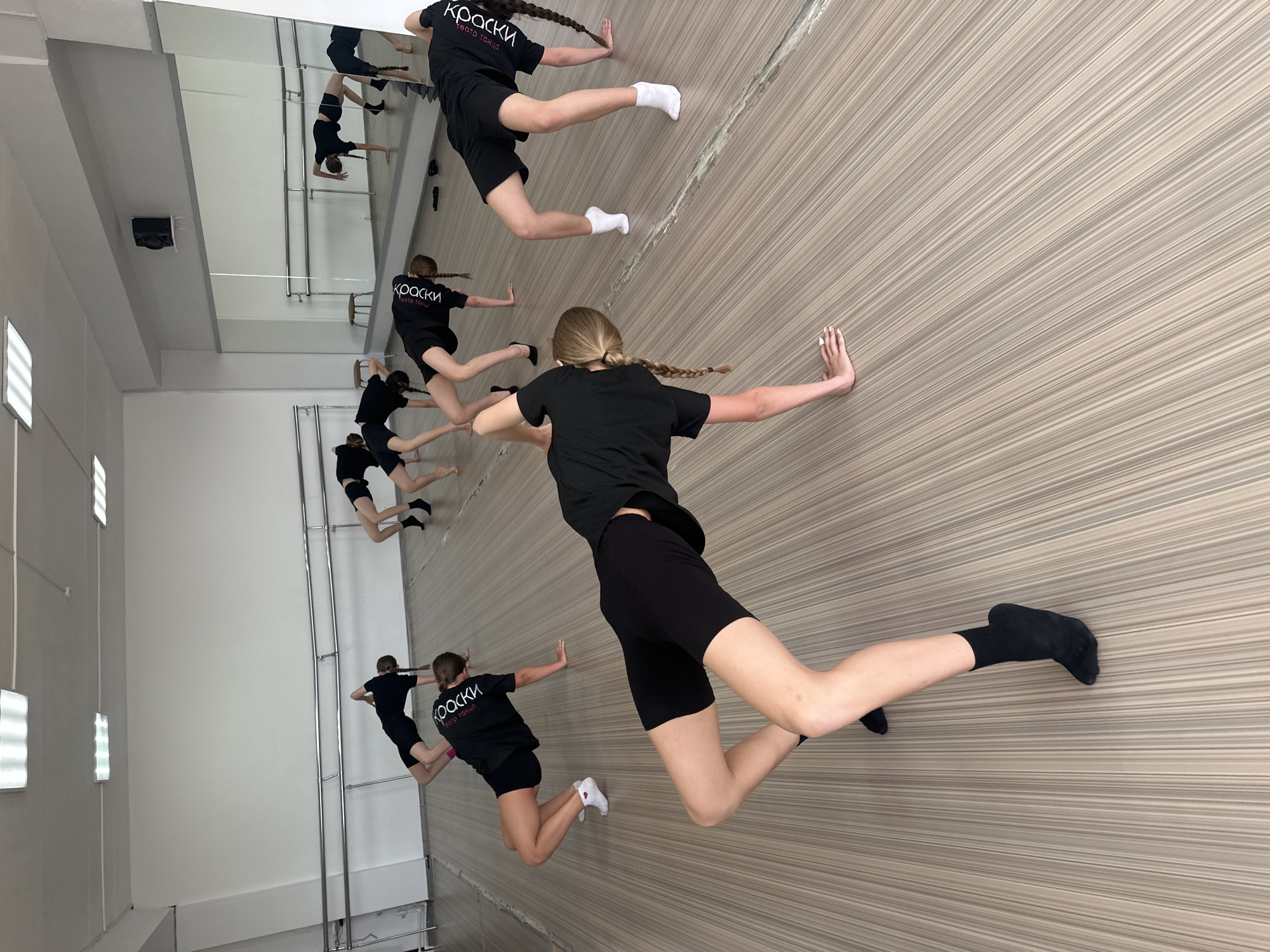 The image shows a dance studio in Votkinsk, Russia, with a mirrored wall reflecting dancers. The primary subject is a young woman mid-movement, wearing a black t-shirt with Cyrillic text "Краски театр танца" (Colors Theater Dance), and black shorts, her blonde hair in a braid. She is extending her right arm to the beige striped wall for balance while balancing on one leg. Other dancers are visible, also wearing the same black attire, performing similar exercises near ballet barres, with their reflections visible in the mirror. The room has white walls and light fixtures.