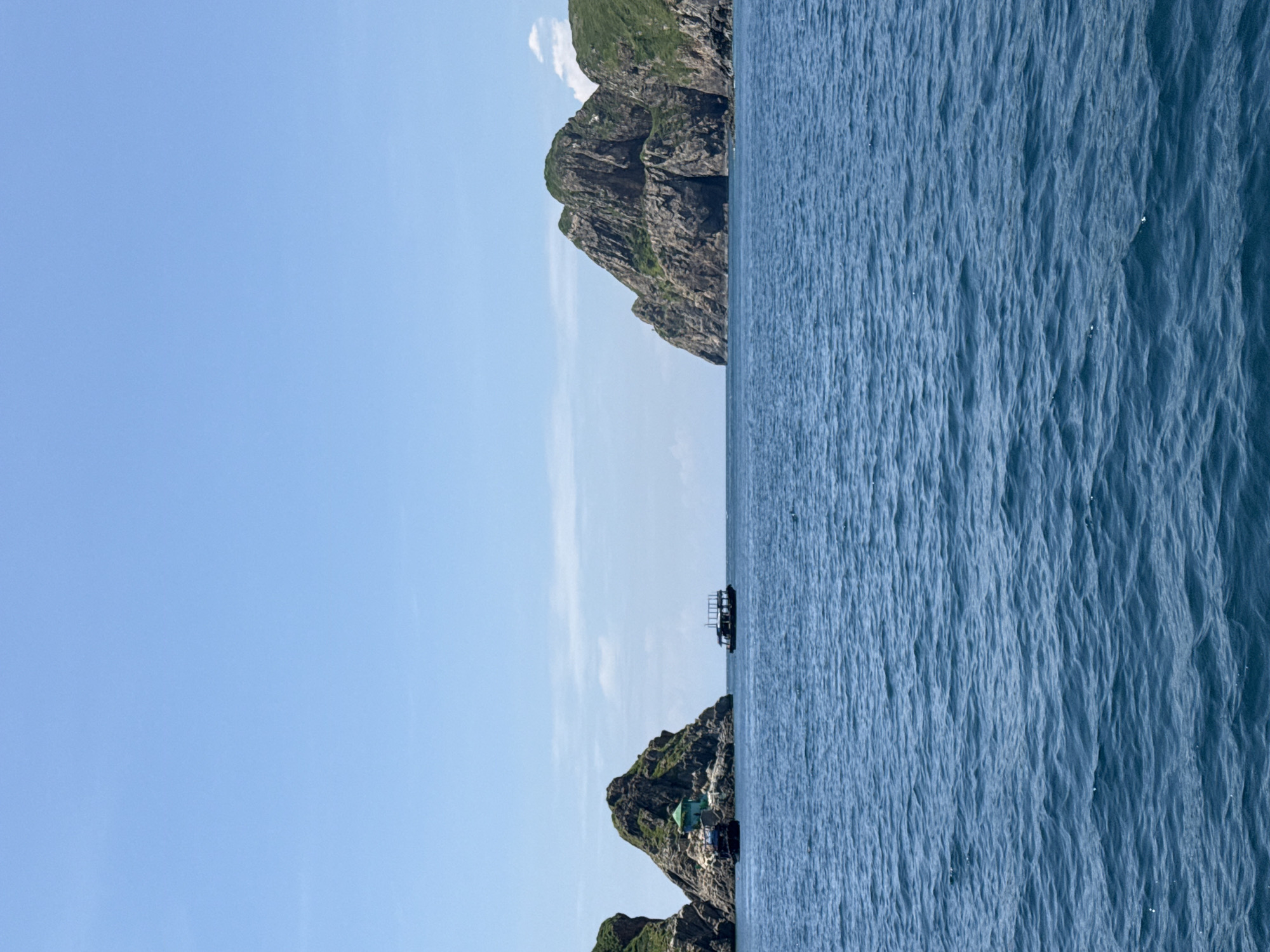 This is a photograph taken from a boat on a bright, clear day, looking out at a body of water with islands and boats. The sea is a deep blue with small waves. In the middle ground, two rocky islands are visible. The island on the left is smaller and has a small green structure on top. A dark-colored boat is moored to its side. Further to the right, a larger, more rugged island with greenery on its slopes rises from the water. Between these two islands, a medium-sized boat with a dark roofed structure is visible. On the right side of the image, a third, larger island dominates the horizon, with steep, rocky cliffs and patches of green vegetation. The sky is a pale blue with a few wispy white clouds. The scene is likely a coastal area or a bay, with the islands forming a natural landscape within the water. Given the presence of similar geographical features and boat types, this scene could be located in the coastal waters around Nha Trang, Vietnam, potentially near Hon Mun Island, known for its marine protected area and rocky outcrops.
