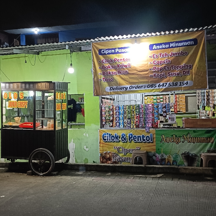 The image shows a street food stall at night, located in Tugu, Indonesia, at coordinates -6.975 latitude, 110.332 longitude. The stall is a small, wooden structure with a black and green facade. The front of the stall features a glass display case that holds various foods, including fried mushrooms, which are labeled “Olos Jamur Crispy.” The stall also has a large sign with an inscription “Cipen Pasan” listing the various food options available. To the right of the stall is another sign with an inscription “Aneka Minuman” that lists the different drinks available for purchase. The stall is lit by several lightbulbs, casting a warm glow on the scene. The ground in front of the stall is paved with gray concrete blocks, and the surrounding environment is dark and quiet. The stall appears to be closed for the night.