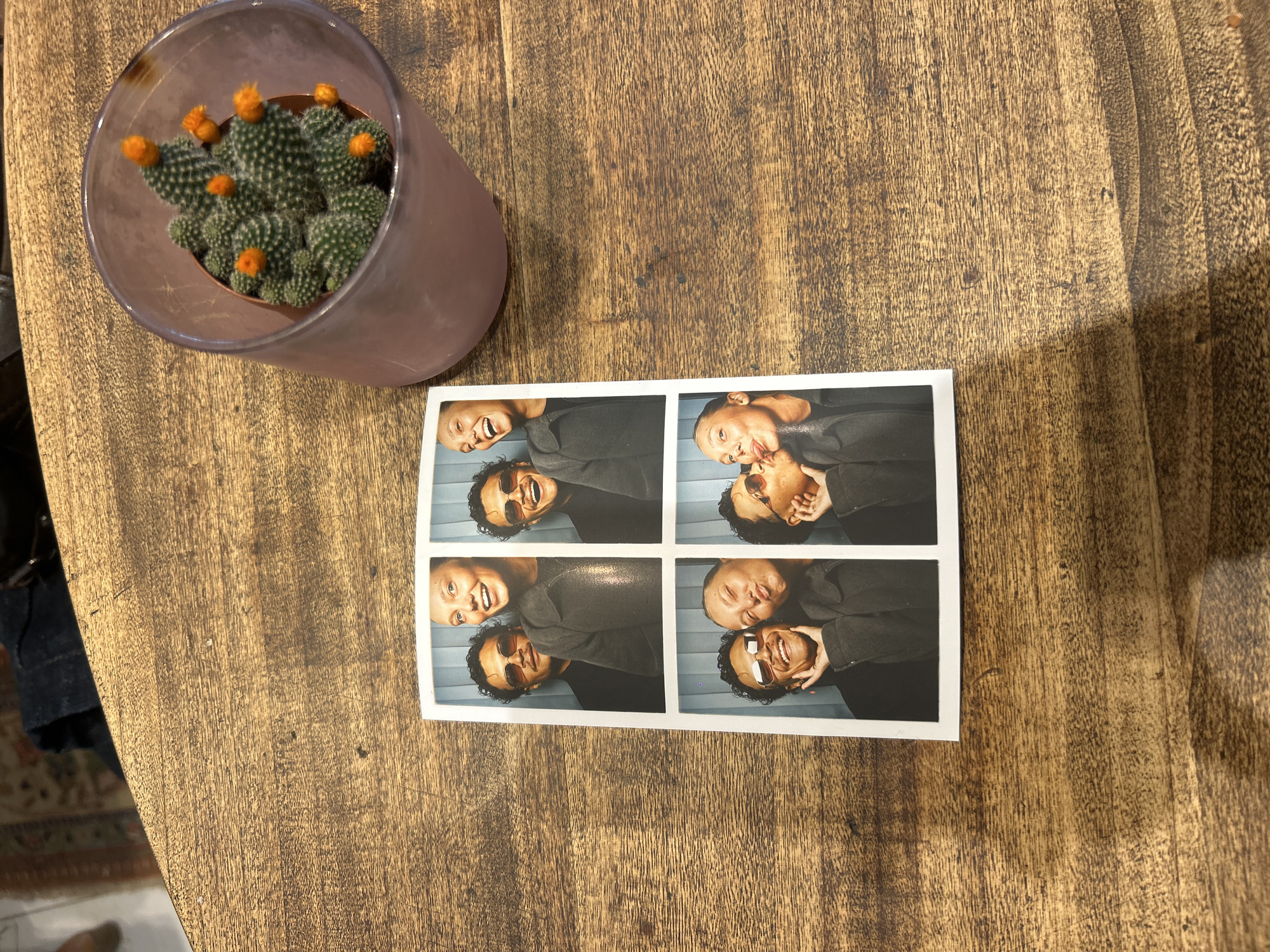 The image depicts a wooden table with a cactus in a frosted glass pot on the left, and a photobooth strip showing four images of a couple on the right. The photos feature a woman with blonde hair and a man with curly dark hair and sunglasses, posing playfully. The background of the photos appears to be a light blue wall.