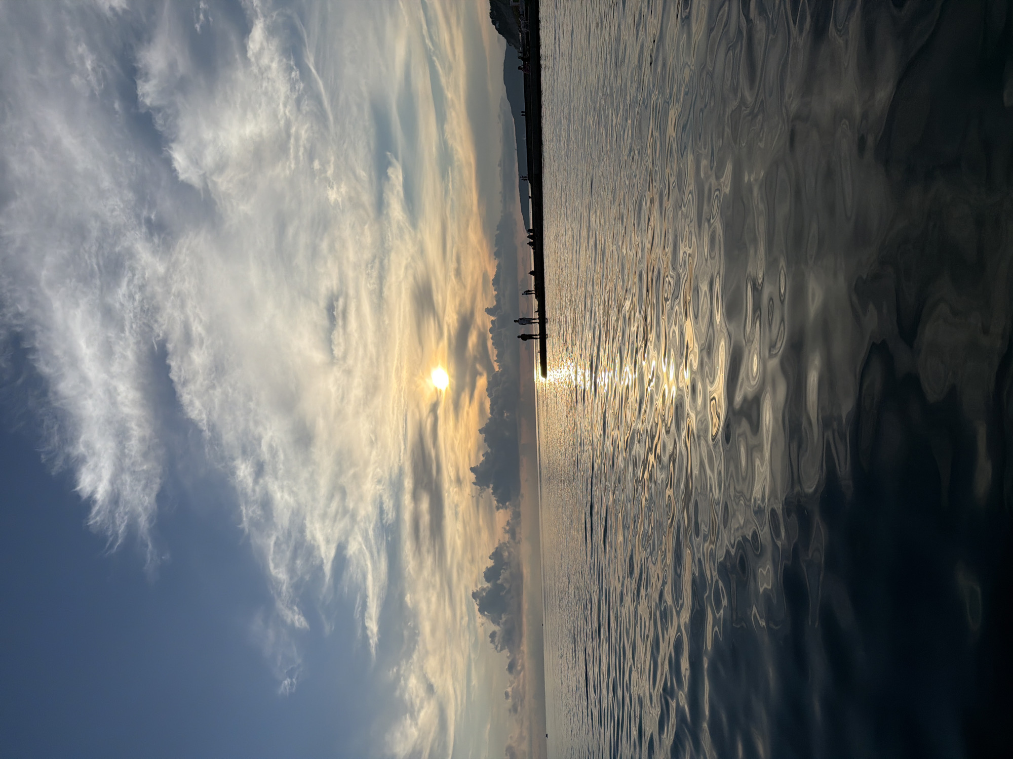 A sunset scene is depicted over the Black Sea from the embankment of Dederkoy, Russia. The sun, a bright orb, is low on the horizon, casting a golden reflection on the rippling water. Wispy clouds streak across a blue sky, some tinged with orange and yellow from the setting sun. Silhouetted figures stand on a concrete pier extending into the sea, appearing to observe the sunset or fish. A line of darker clouds clusters below the horizon, adding depth to the sky. The water is calm, with gentle waves reflecting the colors of the sky.