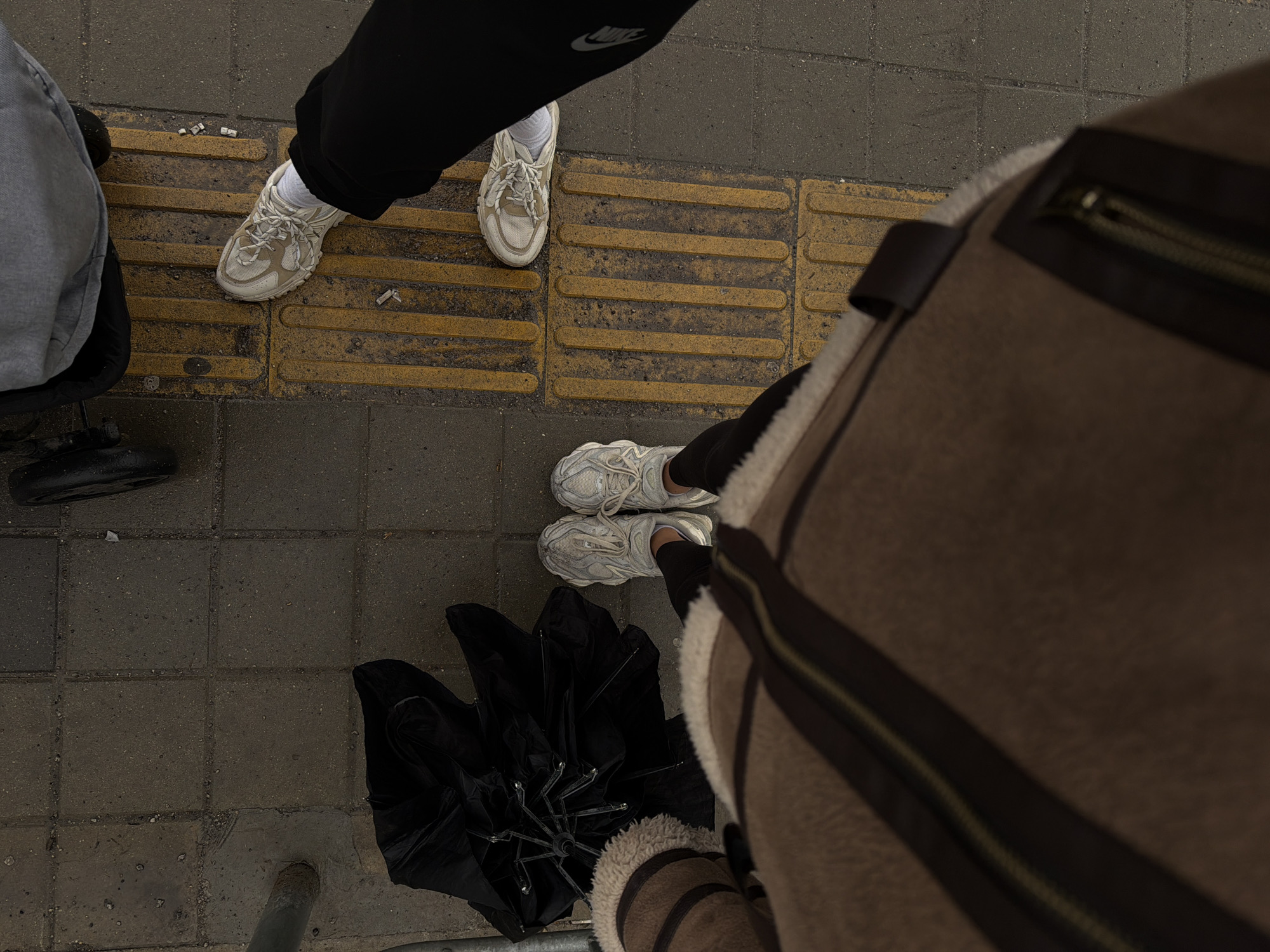 An overhead view captures a segment of an urban sidewalk in Sofia, Bulgaria. The scene shows the lower extremities of two individuals and part of a third subject. In the foreground, a person is observed wearing a brown shearling-lined coat, black pants, and light grey chunky sneakers, standing on dark grey concrete pavers. To their immediate left, a collapsed black umbrella rests on the same paving, near a metallic pole partially visible on the far left edge. Adjacent to this, a second individual's right leg and foot are visible, wearing black pants with a white Nike logo near the ankle and light beige chunky sneakers, positioned atop a section of yellow tactile paving marked with parallel raised strips. Scattered cigarette butts are present on this tactile paving. In the upper right corner, a grey fabric portion and a black wheel of a stroller are discernible. The paving surfaces exhibit signs of wear, and some areas appear damp.
