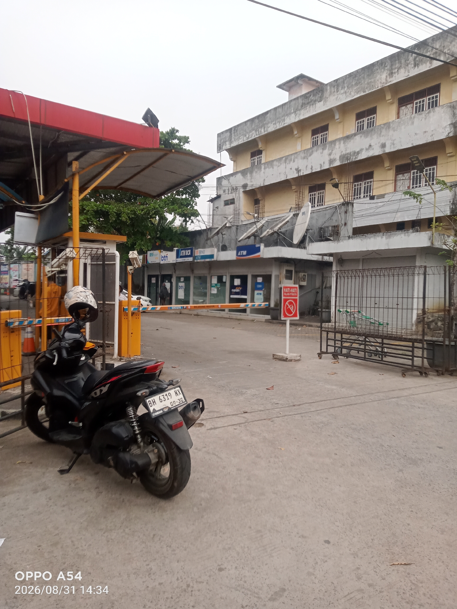 The image captures an outdoor scene in Jambi, Indonesia, featuring a parking area and a multi-story building. In the foreground, a black scooter is parked on a concrete surface. The scooter has a license plate that reads "BH 6319 NX" and "09-30". A white helmet rests on the handlebars. To the left, a red and yellow automated gate structure is visible, with a person in an orange uniform standing behind it.
Behind the gate, a row of shops or offices line the ground floor of a cream-colored, multi-story building. Several signs are visible on these establishments, including "BSI", "BTN", and "ATM BCA". Further back, the building appears aged, with weathered concrete facades and multiple rows of windows. In front of the building, a red and white sign with a red circle and slash over a cigarette symbol indicates "NO SMOKING".
To the right of the shops, a metal gate encloses a section of the property, and several shopping carts are visible behind it. The overall environment suggests a commercial or mixed-use area, possibly a parking lot or entrance to a business complex. The sky is overcast.