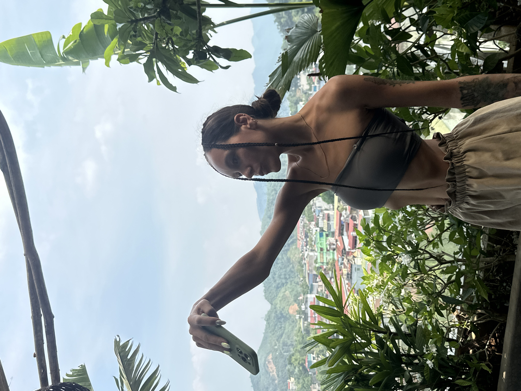 A young woman is captured in a selfie or photo, taken from an elevated viewpoint overlooking a scenic mountainous landscape and a town. The image, likely intended for portrait orientation, shows the woman positioned on the right side of the frame, holding a light green smartphone at arm's length to take a picture. Her body is angled slightly, and her head is turned, looking down at the phone's screen with a focused, neutral expression.
She has long, dark hair styled in a bun, with several long, thin braids framing her face. She is wearing a dark gray or green strapless tube top and high-waisted, light beige or khaki ruffled trousers. Visible tattoos adorn her left arm. Her fingernails are long and manicured with a light, possibly iridescent, polish.
The background reveals a expansive vista under a clear, bright daytime sky. Lush green mountains, densely covered in vegetation, stretch across the horizon. A town or settlement is nestled in the valley below, with numerous buildings visible among the greenery, some featuring colorful roofs. Various large, vibrant green leaves and plant stalks, including some banana-like foliage and smaller plants with white flowers, frame the scene, suggesting a tropical or subtropical environment. The overall scene, with its dense green mountains and valley town, is consistent with the provided location context of Hà Giang 1 Ward, Vietnam. No discernible text is visible in the image.