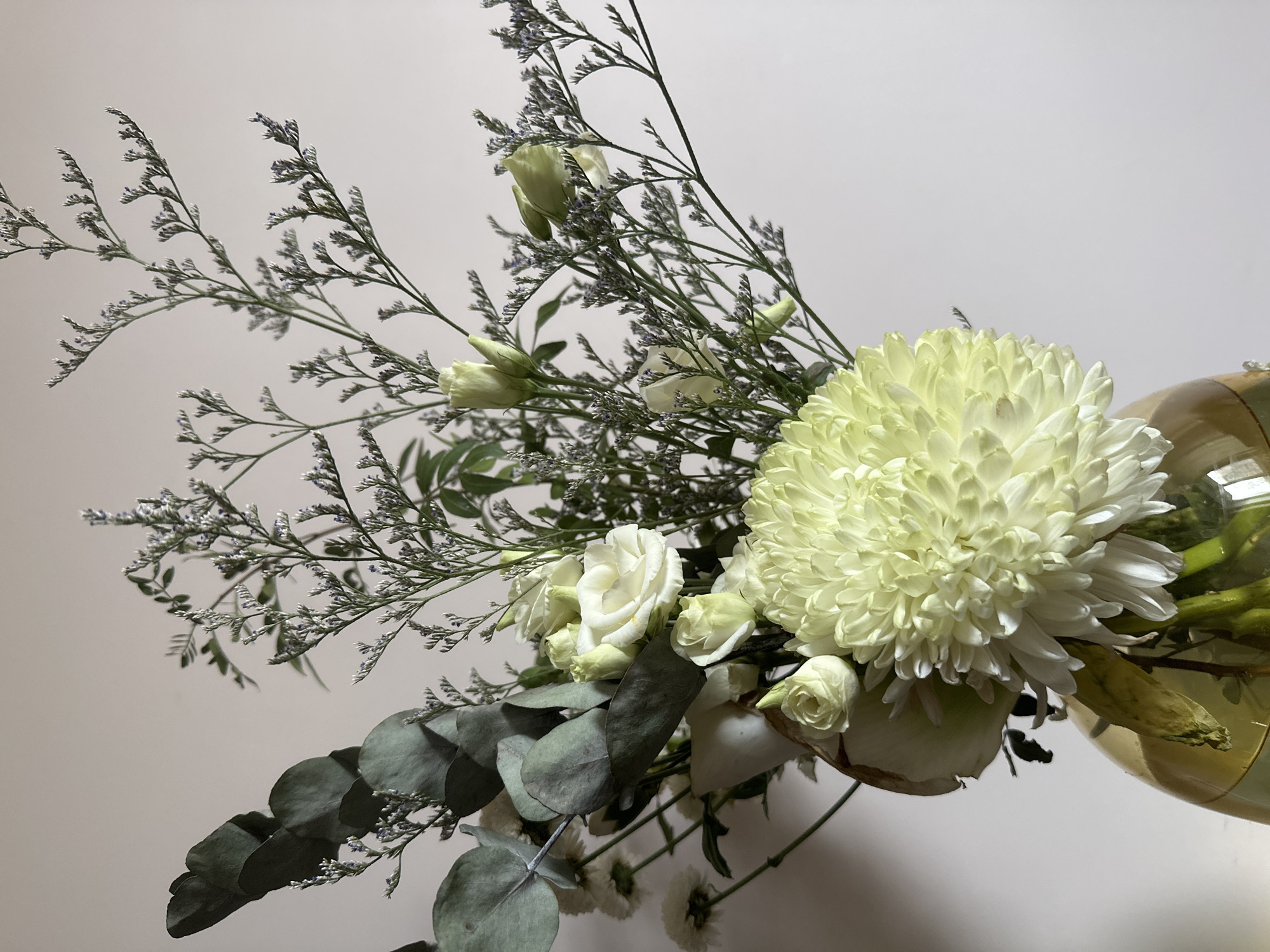 A vase of flowers, including a large white chrysanthemum, smaller white roses, eucalyptus leaves, and other assorted greenery, sits against a plain white background. The vase is made of glass and appears to have a yellowish tint. The composition is slightly angled, giving a view from the bottom up. The lighting is diffuse and even, revealing the textures of the flowers and leaves. There is no indication of the city of Krasnoyarsk, Russia.