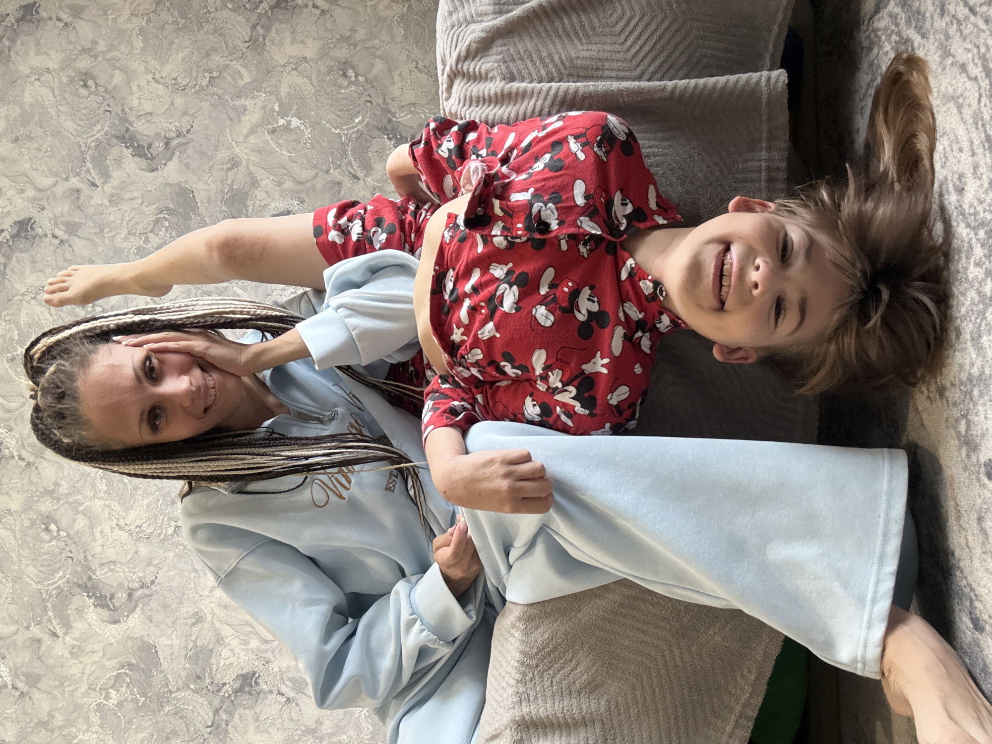 An indoor portrait features two smiling individuals, an adult woman and a child, lounging on a gray, textured sofa or bed. The image is taken from a slightly overhead perspective, with a light gray marbled wall visible in the background.
The woman, positioned on the left side of the frame, has long, multicolored dreadlocks or braids. She is wearing a light blue zip-up hoodie with stylized dark text embroidered on the front, which appears to read "Vintage." Her hand is near her face, and she gazes towards the camera with a warm smile.
The child, on the right side of the frame, is wearing bright red pajamas adorned with numerous white Mickey Mouse patterns. The child has light brown, somewhat tousled hair and is smiling widely, showing teeth. One of the child's bare feet is visible near the woman's head. The woman's arm appears to be resting near the child, suggesting closeness and affection.
Both subjects display joyful expressions, contributing to a happy and relaxed atmosphere. The casual clothing, especially the child's pajamas, indicates a comfortable, leisurely moment, possibly in the morning or evening within a home setting in Rusanovo, Russia.