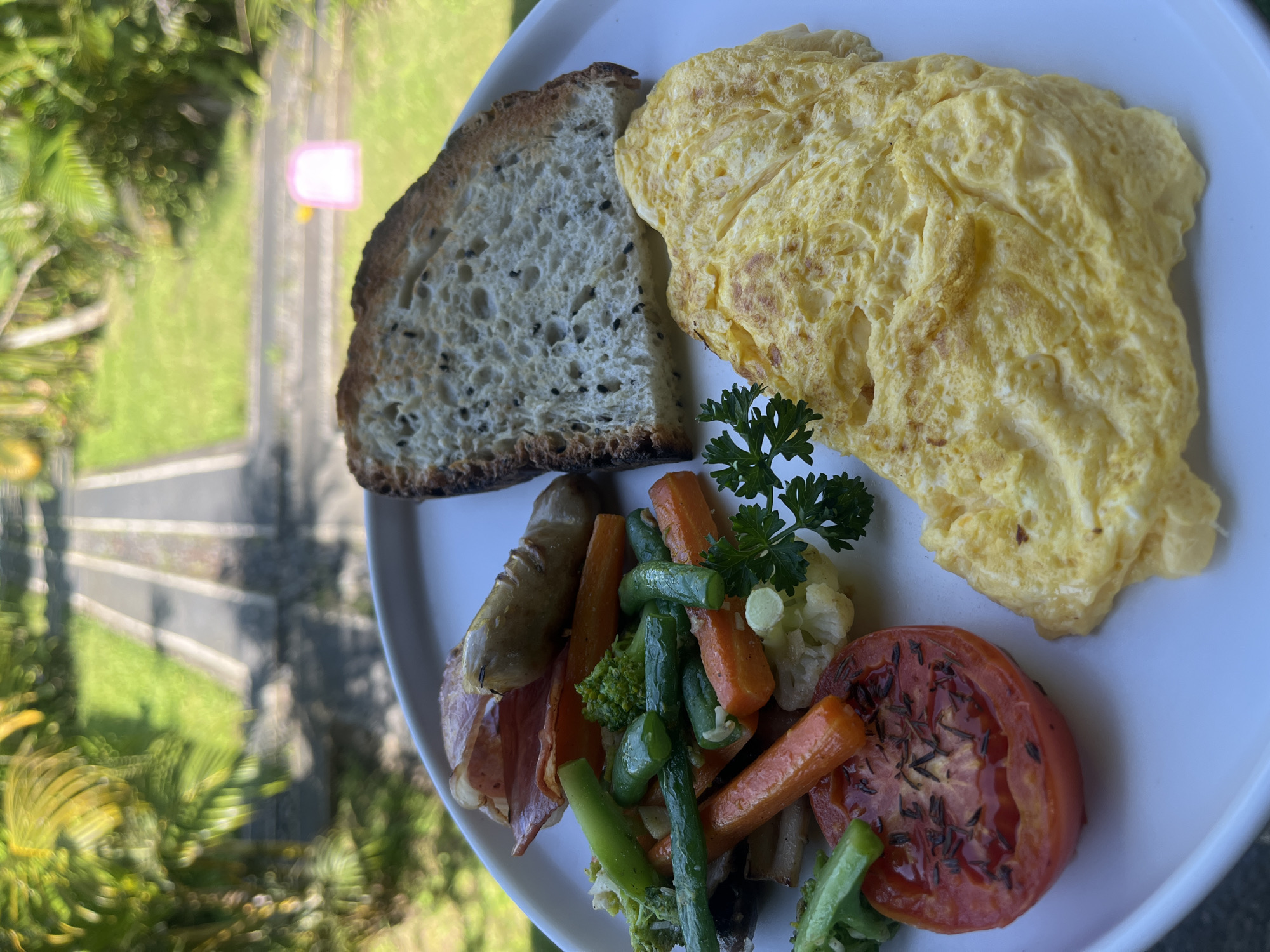 A close-up, overhead shot displays a white plate filled with breakfast items, set against a blurred background of tropical foliage and a paved path. On the plate, a golden-yellow omelet is folded and placed prominently in the foreground. To its right, a slice of toasted bread with visible seeds is positioned. The left side of the plate features a colorful medley of vegetables, including bright red tomato slices with herbs, green beans, carrots, broccoli florets, and cauliflower. Behind the vegetables, a piece of cured meat and a sausage link are visible. A sprig of parsley garnishes the vegetable portion. The environment suggests an outdoor setting, possibly a restaurant or resort in Tegallalang, Indonesia, given the lush greenery and the distinct path.