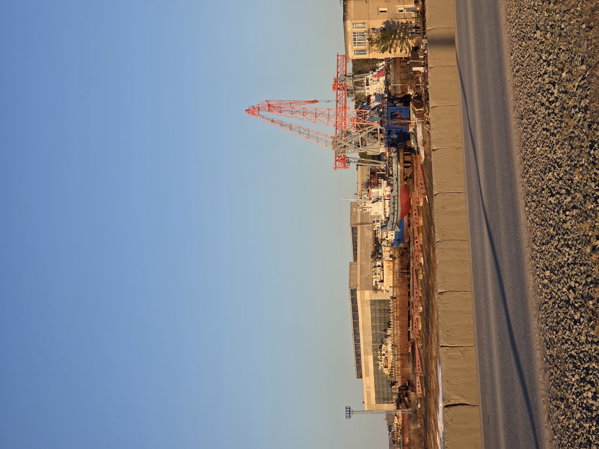 The image depicts the Kama shipyard, located in the city of Chaikovsky, Russia. A large, red gantry crane is visible in the foreground, towering over the shipyard's infrastructure and vessels under construction or repair. Several buildings, including residential apartments, are visible near the waterfront. A road separates the shipyard from an area covered with gravel. The sky is a clear blue.
