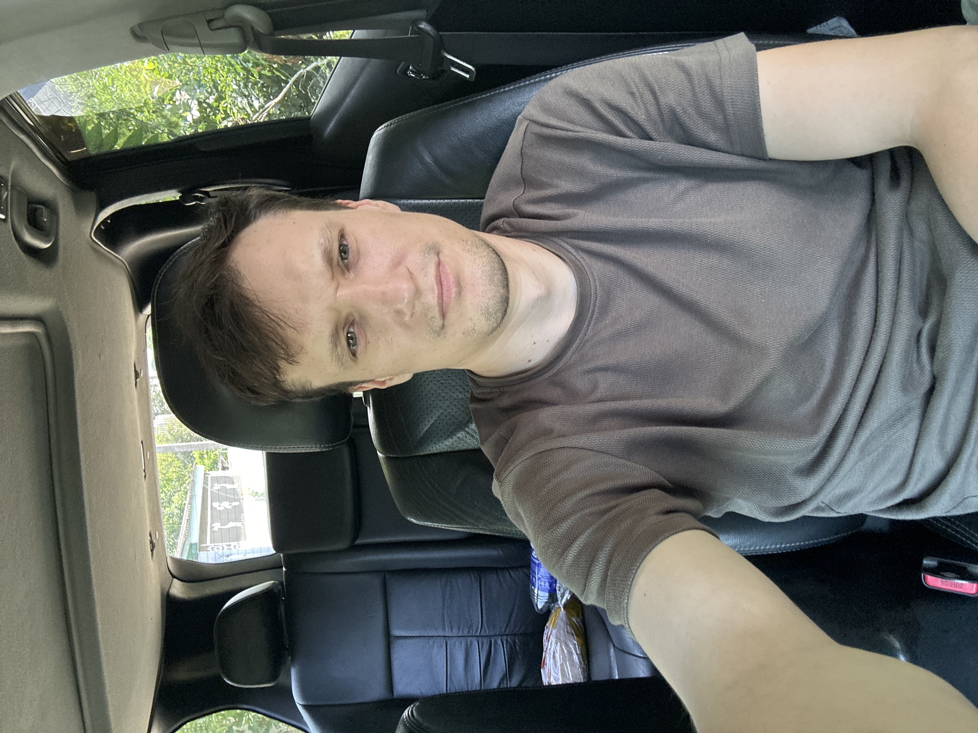 A man is sitting in the driver's seat of a vehicle. He is looking at the camera and appears to be taking a selfie. The interior of the vehicle is visible, including the black leather seats and the ceiling. Outside the vehicle, greenery and a building can be seen through the windows. The man is wearing a brown t-shirt. There are some items on the back seat, including a plastic bottle and a package. The scene suggests the man is in a parked vehicle.