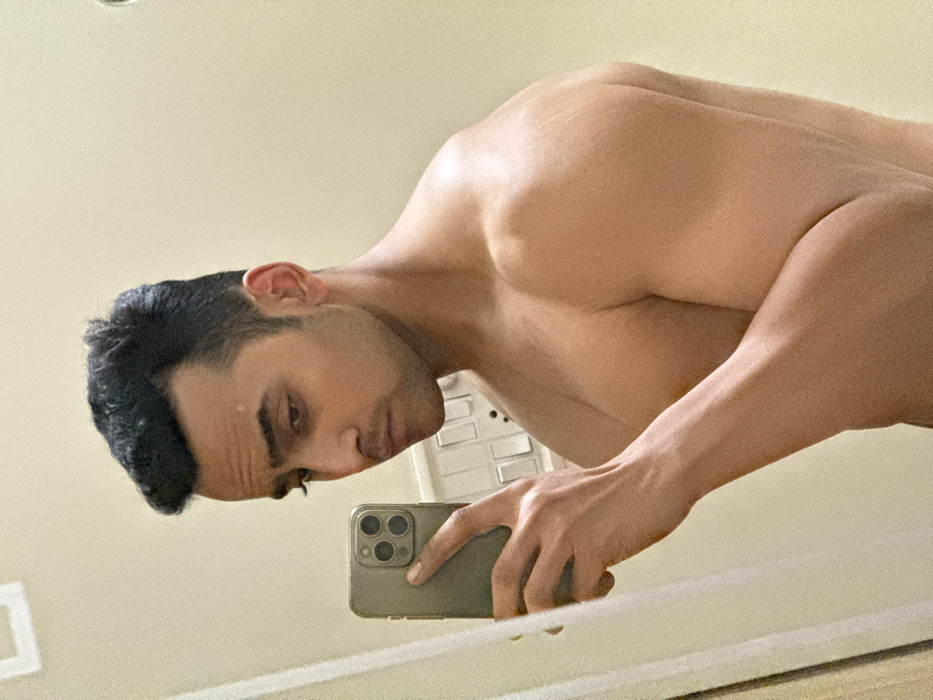 A muscular, shirtless man is seen taking a selfie in a mirror. His head is tilted down as he looks into his phone, which he holds in his right hand. The phone has three camera lenses. The man has short, dark hair and appears to be well-groomed. In the background, a light switch is visible on the wall. The wall itself is a pale color, suggesting a clean and simple interior.