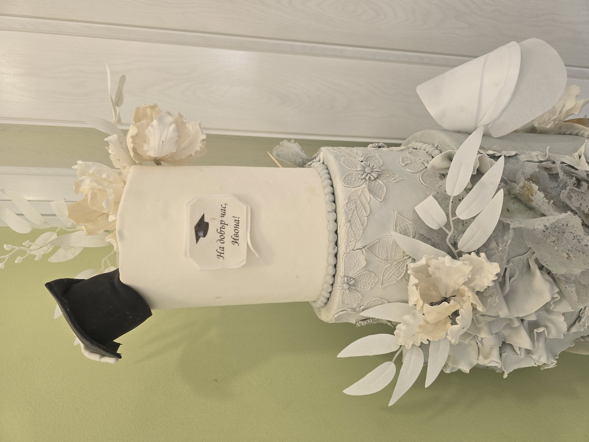 A three-tiered cake, decorated with white fondant flowers and foliage, is set against a pale green wall and white wooden doors. The top tier features a black graduation cap and a label with text and an image of a graduation cap. The middle tier has a textured lace pattern. The bottom tier is adorned with ruffles and more white flowers and foliage. The cake appears to be a celebratory item, possibly for a graduation. The image does not contain any information to suggest a specific location in Dobrich, Bulgaria.