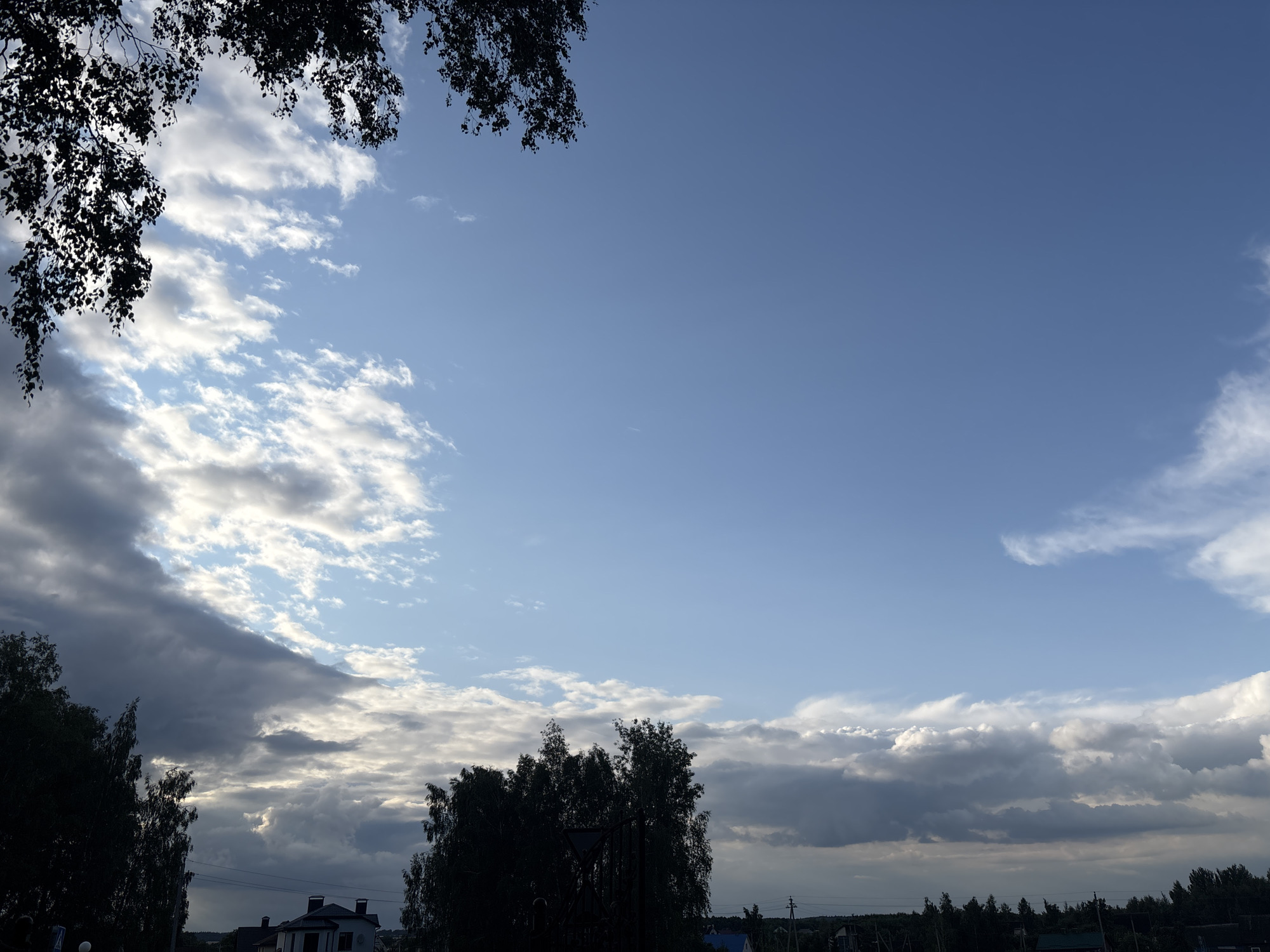The image depicts a wide shot of the sky over Kapyĺ, Belarus, with a low-lying landscape visible at the bottom. The sky is a mix of blue and dramatic clouds, with patches of sunlight breaking through darker storm clouds. Silhouetted trees frame the left and right edges of the scene, and the tops of some buildings, possibly houses, are visible on the horizon, along with power lines and a dense line of trees. The overall impression is one of an approaching or receding storm under a vast, partly cloudy sky.
