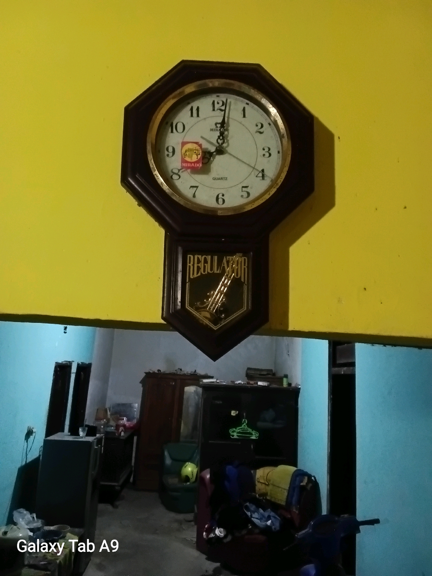 A wall clock with a dark brown octagonal frame hangs on a yellow wall. The clock face is white with black numbers and hands. A small red and gold sticker with the word "MIRADO" is visible on the clock face. Below the clock face is a dark brown pendulum casing with the word "REGULATOR" in gold lettering. The clock appears to be functional. The image also shows a room with blue and light blue walls, a dark brown cabinet, a dark gray refrigerator, a green chair, a motorcycle, and various objects on the floor and shelves. The overall impression is a cluttered but lived-in space.