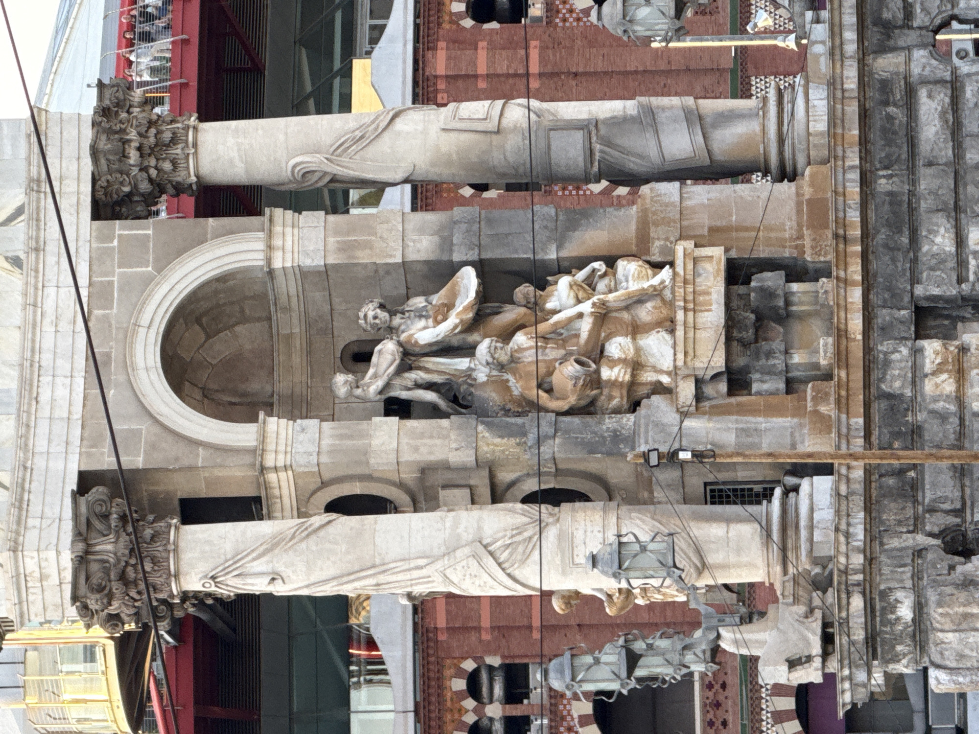 The image depicts the Monument to Columbus (Monumento a Colón) in Barcelona, Spain. This monument is located at the end of Las Ramblas, near Port Vell. The central structure is a tall, fluted column topped with a statue of Christopher Columbus pointing towards the sea. The base of the monument features a circular pedestal with allegorical sculptures and various architectural elements. In this particular view, the focus is on the lower portion of the monument, showcasing several stone sculptures of figures in classical attire. There are also decorative architectural details, including columns, arches, and ornate capitals. In the background, modern buildings and structures are visible, indicating an urban setting. The lighting suggests it is daytime, with shadows cast by the architectural features and sculptures.