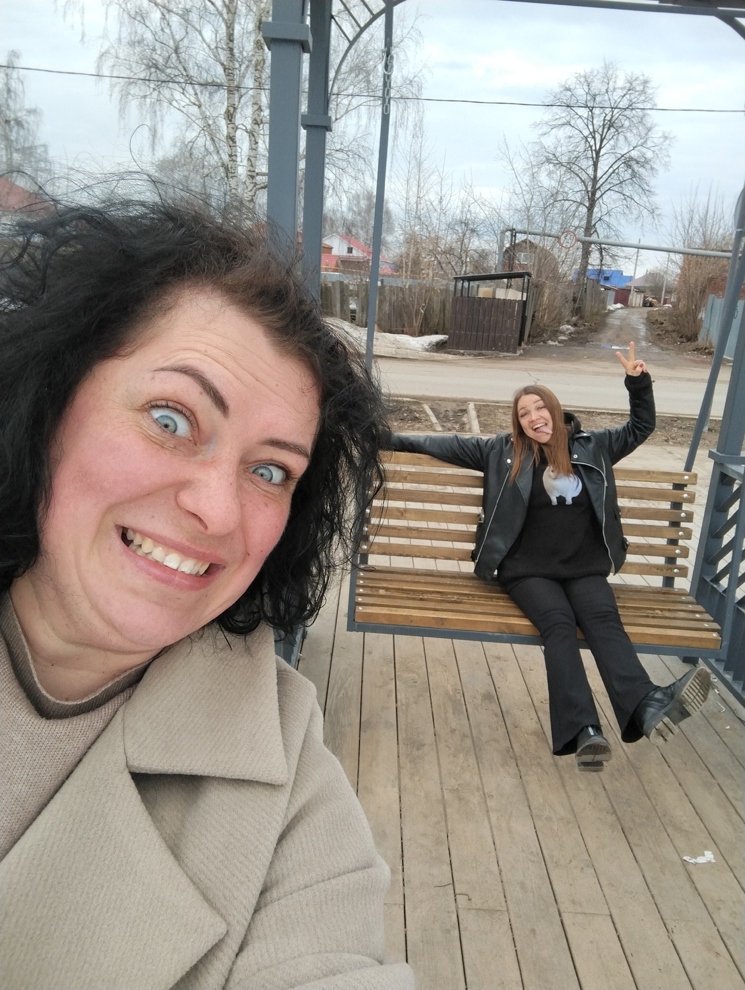 Two women are posing for a photo on an outdoor swing in Votkinsk, Russia. The woman in the foreground has dark, messy hair and is wearing a beige coat. She is smiling widely and making a surprised expression with her eyes. The woman in the background is sitting on the swing, wearing a black leather jacket, black sweater and black pants. She is holding up two fingers in a peace sign. The background includes a street with houses and bare trees, consistent with late winter or early spring in Russia.